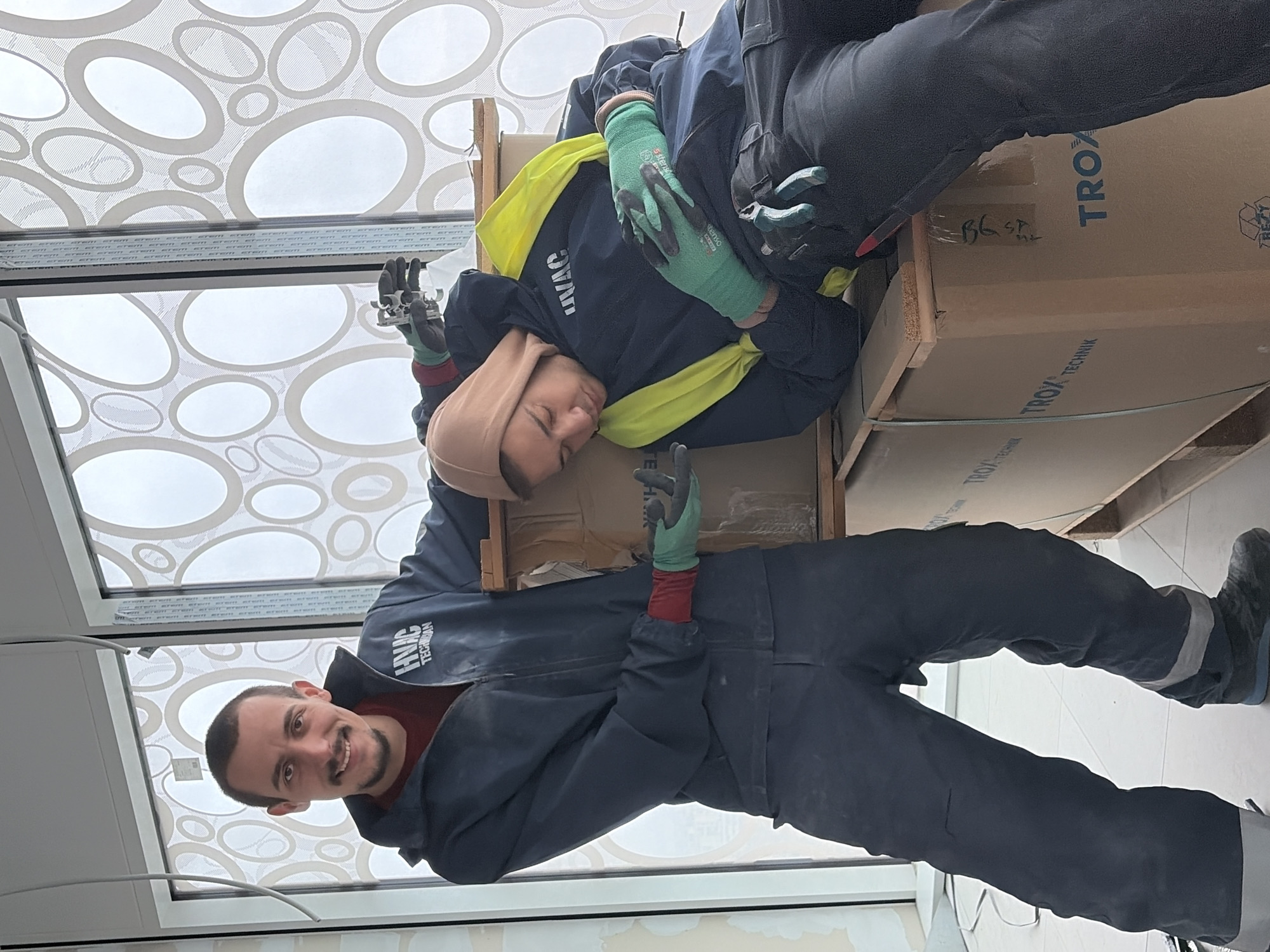Two people are in the photo. One man stands and looks directly at the camera, smiling. He wears dark blue overalls with the inscription "HVAC TECHNICIAN" on the chest, a red shirt beneath, and green and red gloves. The second person is lying on boxes and wearing a brown head covering, a dark blue jacket with a yellow vest, and green gloves. The boxes have the word "TROX TECHNIK" printed on them. The location appears to be inside a building, possibly under construction or renovation, with a decorative window with patterned circles in the background. The location is not specified to be in Sofia, Bulgaria.