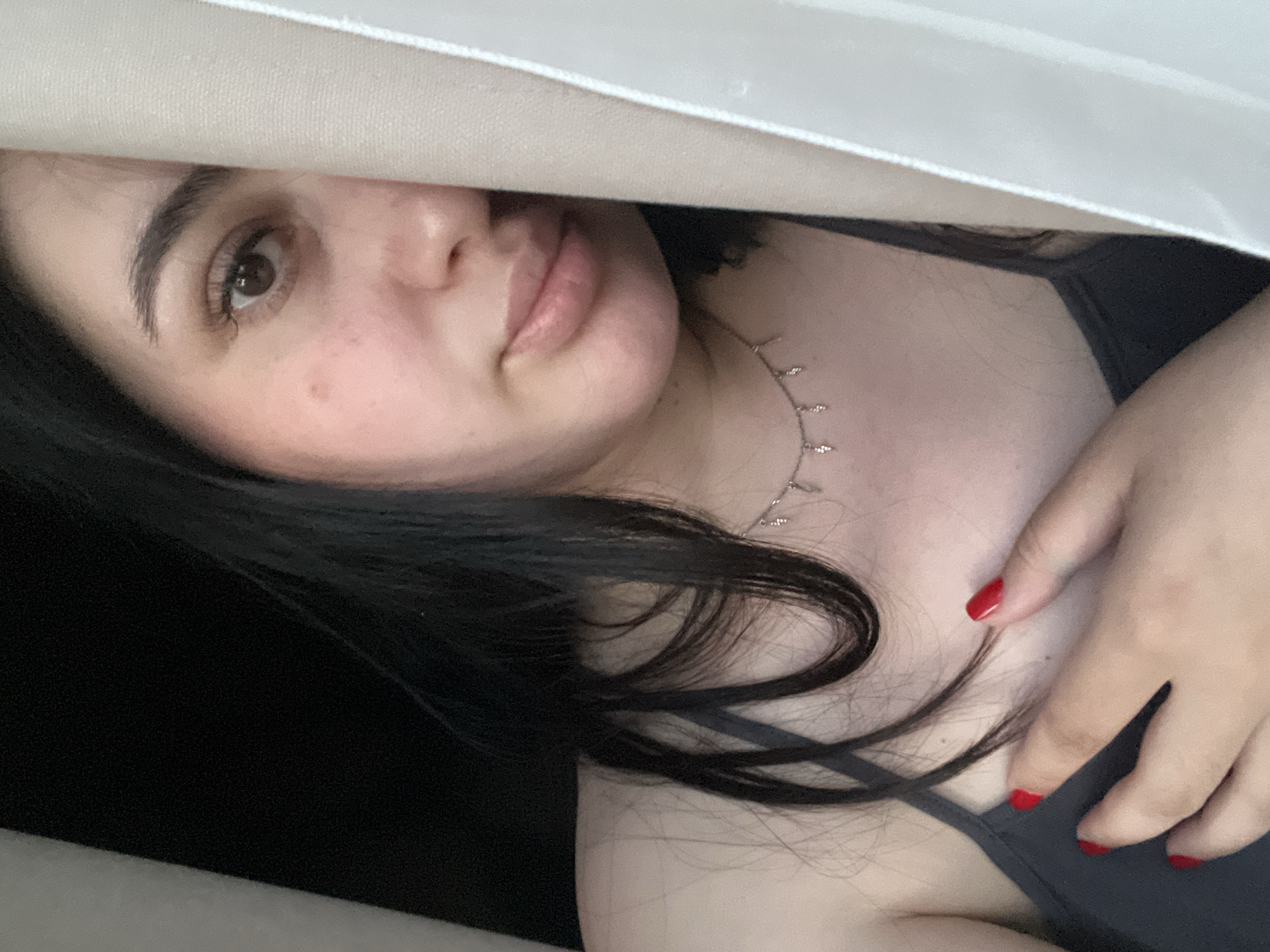 A young woman is posing for a selfie. She has long, dark hair and fair skin. She is wearing a gray tank top and a delicate silver necklace. Her fingernails are painted red. The woman is partially obscured by a white curtain. The background is dark, and the location is unspecified, though Chaikovsky, Russia, is mentioned.