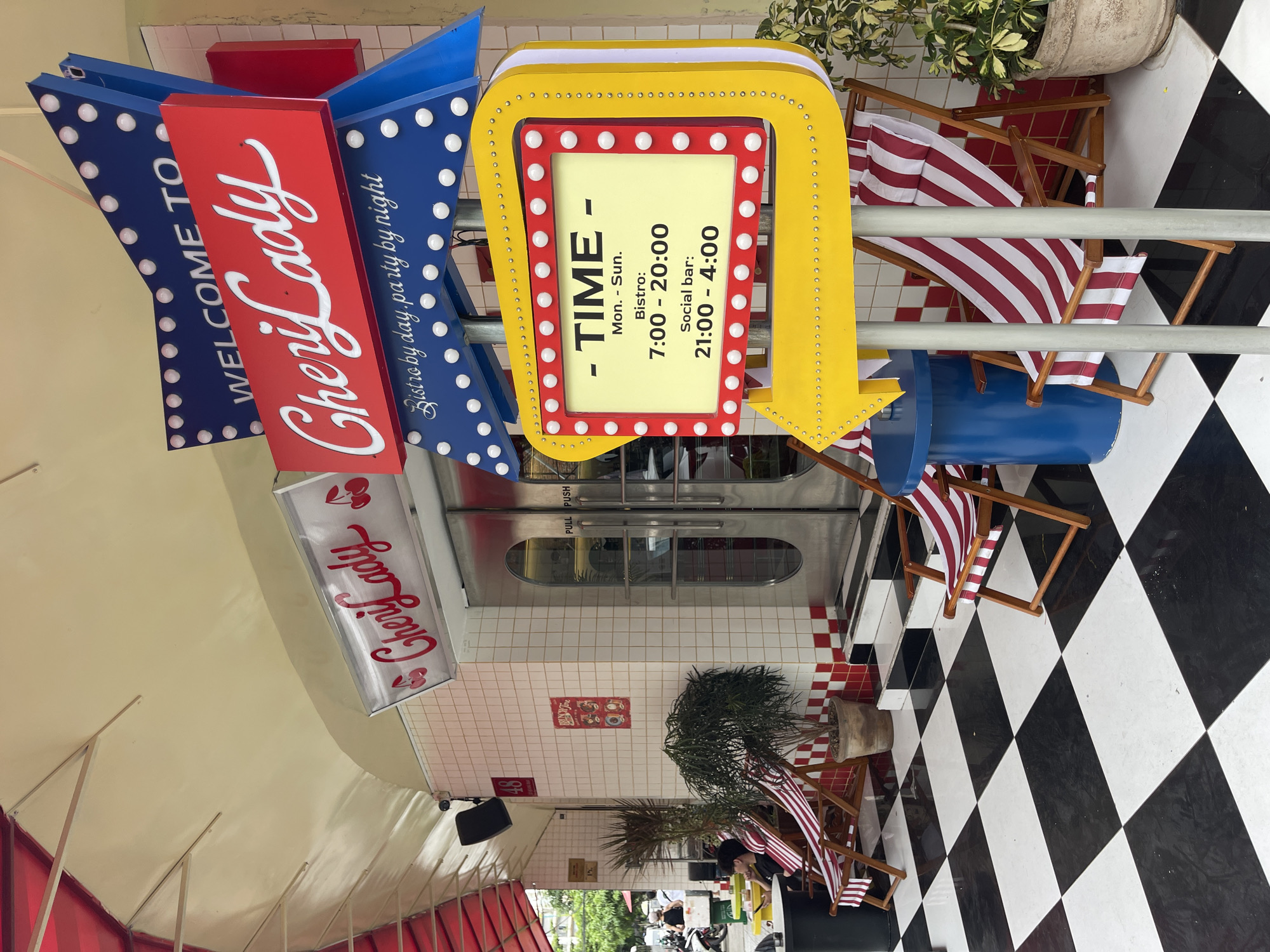The image depicts the exterior of a establishment named "Cheri Lady" in the city of Da Nang, Vietnam. The facade features a tiled wall and a glass door with "PULL PUSH" signage. In front of the establishment, a checkered black and white tiled floor is visible, along with two striped deck chairs and a blue side table.
Dominating the scene is a large, retro-style sign with "WELCOME TO Cheri Lady" prominently displayed. Below this, smaller text reads "Bistro by day, party by night." Another illuminated sign shaped like a directional arrow displays operating hours: "Mon. - Sun. Bistro: 7:00 - 20:00, Social bar: 21:00 - 4:00".
Several people are visible in the background, including individuals on motorcycles and one person sitting on a deck chair. Plants are placed near the entrance, adding to the outdoor seating area. The overall aesthetic suggests a casual, vibrant establishment.