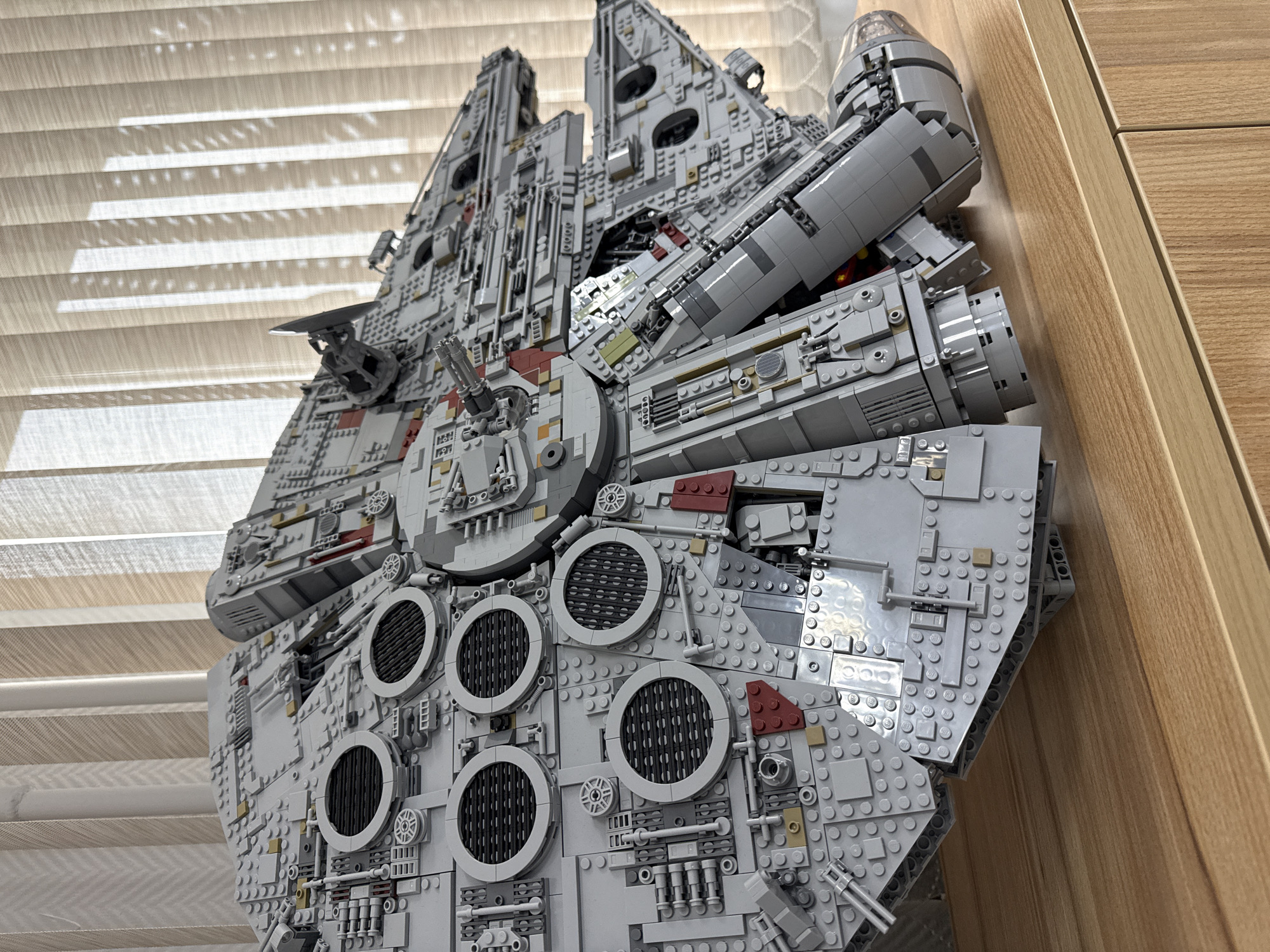 The image shows a LEGO Millennium Falcon model. The model is composed of gray and black LEGO bricks, with detailing to resemble the spacecraft from the Star Wars franchise. It is positioned against a backdrop of closed, horizontal window blinds and a light-colored wooden surface, likely a wall or piece of furniture. The scene appears to be indoors. There is no indication of location in Krasnoyarsk, Russia.