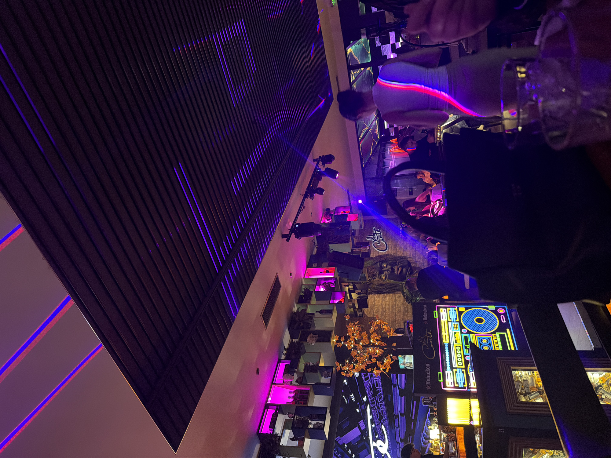 This image depicts the interior of a nightclub or bar, likely in Sofia, Bulgaria, based on signage and design elements. A woman in a white dress stands near a table with glassware. A person, possibly a bartender, is visible behind the counter. The environment is dimly lit, with purple and pink lighting creating a vibrant atmosphere. Decorations include a "Cats" logo, artwork of a man with dreadlocks, shelves filled with objects illuminated in pink, a decorative light fixture resembling a tree with golden leaves, and a colorful neon sign resembling a DJ console. The architecture features dark, angled ceiling panels with embedded purple lights. A Heineken advertisement is also visible.