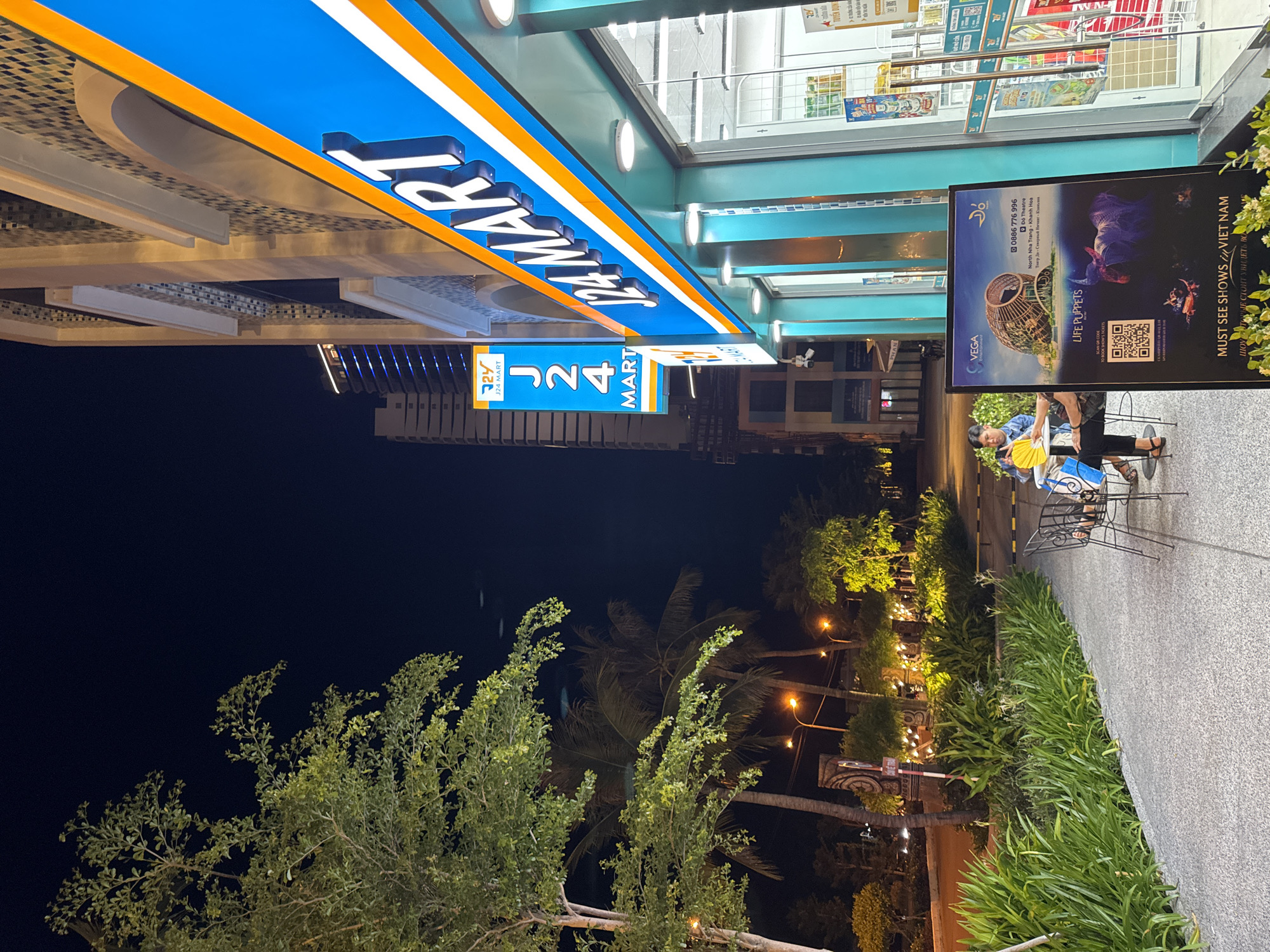 The image shows an exterior view of a "J24 Mart" convenience store at night, located in Bac Nha Trang Ward, Vietnam. The store has a prominent blue and orange sign with illuminated lettering. In front of the store, there is a sidewalk and a landscaped area with lush green plants and trees. To the left of the store, a person is seated at a small table with chairs, holding a yellow fan. A large poster is displayed near the entrance of the store, featuring an image of an elephant and text that includes "LIFE PUPPETS" and "MUST SEE SHOWS // VIET NAM". In the background, a modern high-rise building with blue lighting is visible. Streetlights illuminate the area, casting a warm glow on the scene.