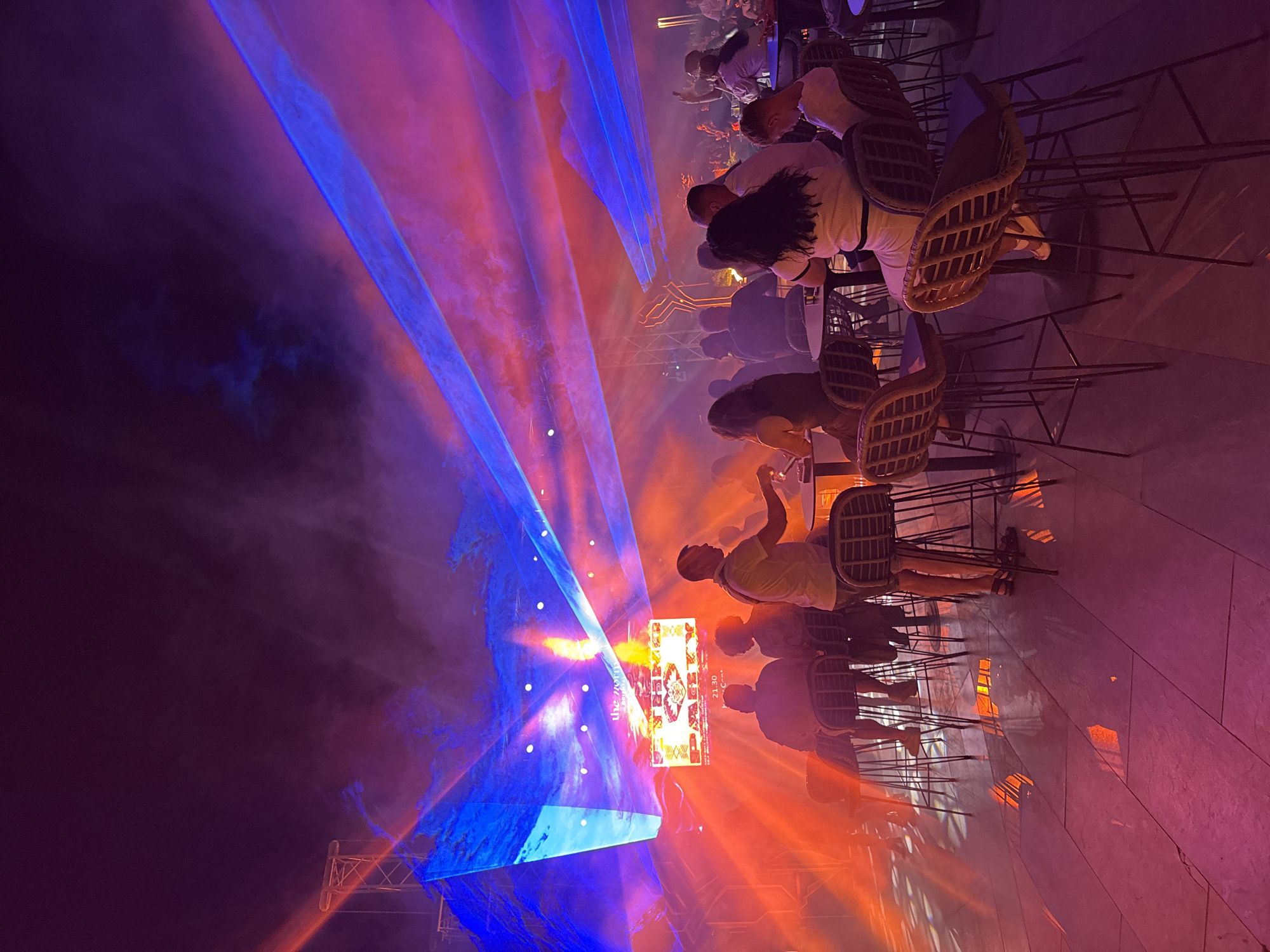The image depicts a vibrant nighttime scene, likely at an outdoor venue in Kemer, Turkey. A laser light show illuminates the sky with streaks of blue and red light, cutting through a smoky atmosphere. In the foreground, several individuals are seated at high-top tables, some appearing to be patrons of a bar or club. One man, wearing a light-colored t-shirt and shorts, stands near a table, holding a drink. The floor is tiled and reflects the colorful lights. In the background, a large screen displays "STONE PAGY" and a time, suggesting an event or performance is taking place. The overall ambiance is festive and energetic, characteristic of a nightlife setting.