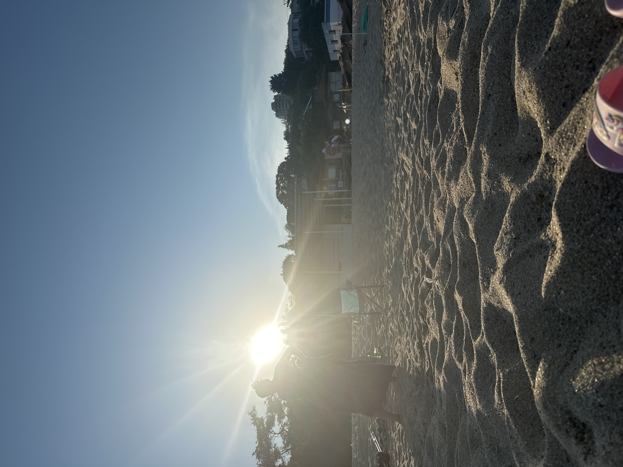 This is a low-angle, full-shot, outdoor photograph of a sandy beach. The sun is low in the sky, casting strong rays and creating lens flare. In the foreground, the rippled sand is visible, with what appears to be a pink and white sandal in the lower right corner. In the mid-ground, a person in a long, dark dress stands with their hands on their hips, silhouetted against the sun. To their right, a folded beach chair with a light blue seat is visible. In the background, there are trees and buildings on a hill overlooking the beach. Several other people are visible in the distance, some walking and some standing near the buildings. The sky is a clear blue with some wispy clouds. The overall impression is a sunny day at the beach.