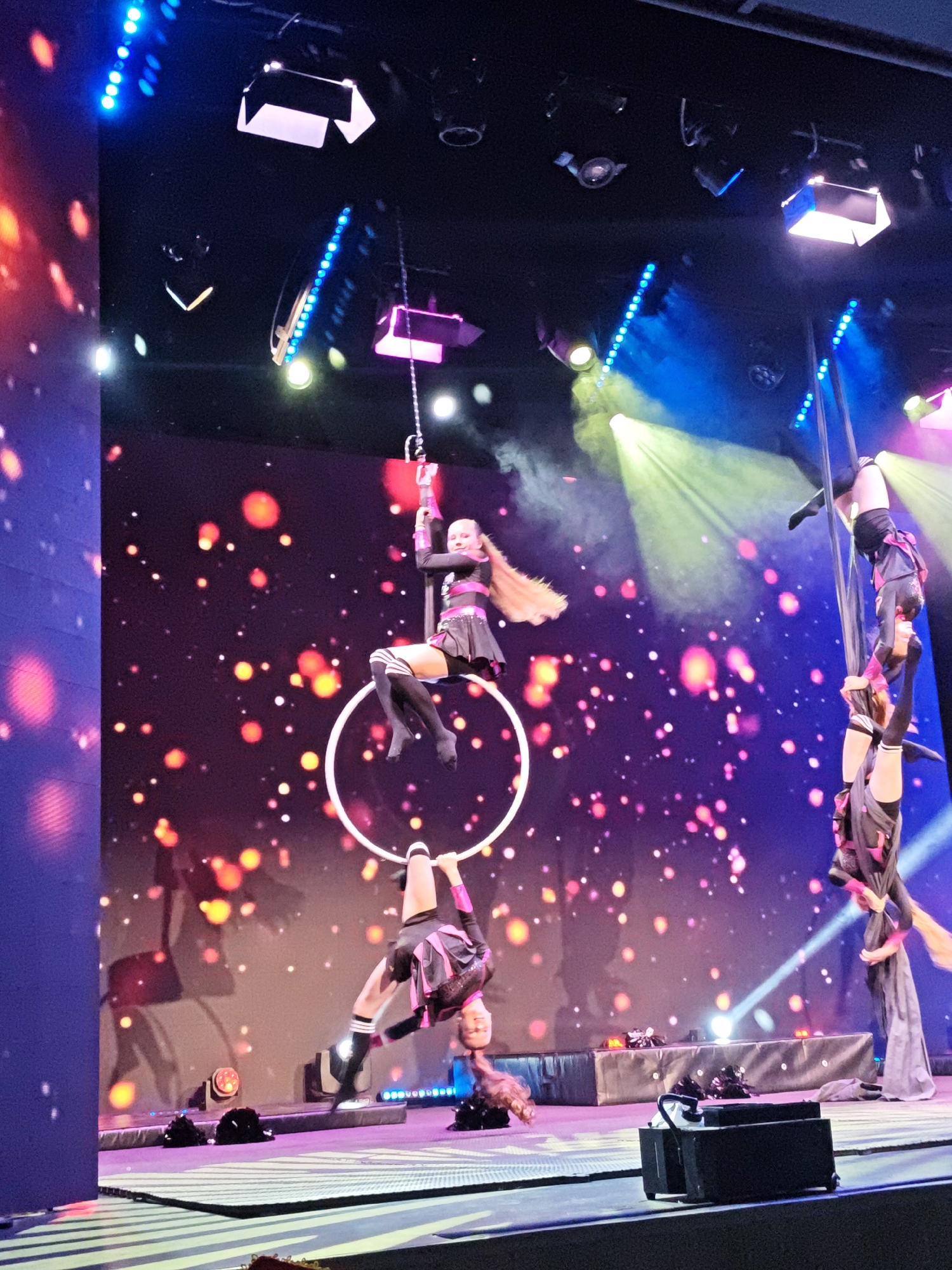 The image depicts a performance art scene, likely a circus act, on a stage under bright, colorful stage lighting. Two women are suspended in the air by a circular apparatus. One is upright while the other is upside down. To the right of them, another group of three women stand vertically, one atop the others’ shoulders. The background is a screen displaying abstract colored lights. The setting is likely a theater or performance venue in Yekaterinburg, Russia.