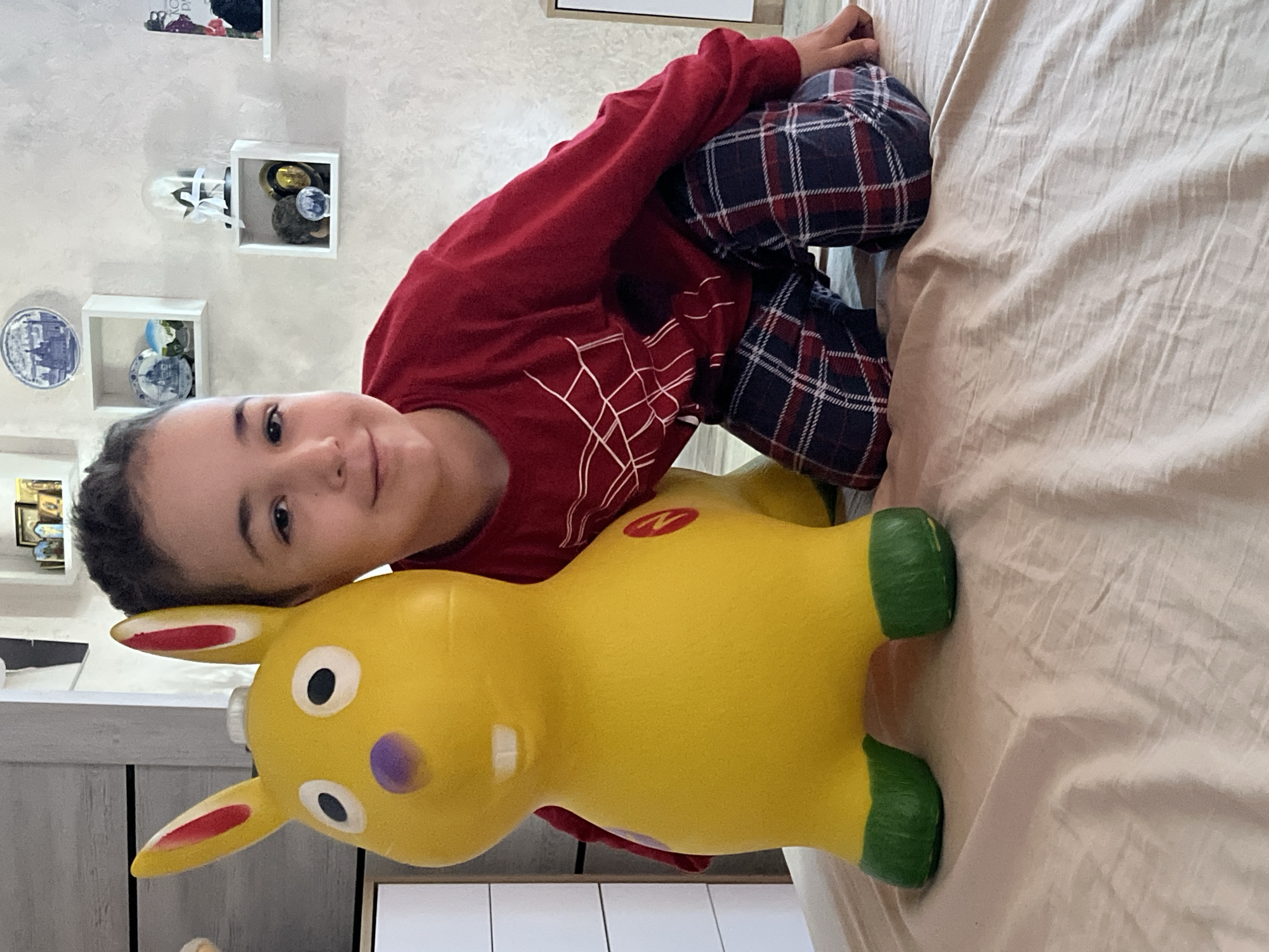 A young boy is kneeling on a bed next to a yellow inflatable rabbit. The boy is wearing a red long-sleeved shirt with a white web-like pattern and plaid pajama pants. The rabbit is bright yellow with green feet, red-lined ears, a purple nose, and two white teeth. The bed is covered with a light-colored sheet. In the background, there are shelves on a textured wall with various decorative items, including plates, figurines, and a book. A white dresser is partially visible on the left. The overall setting appears to be a child's bedroom. The image is taken in the city of Krasnyye Barrikady, Russia.