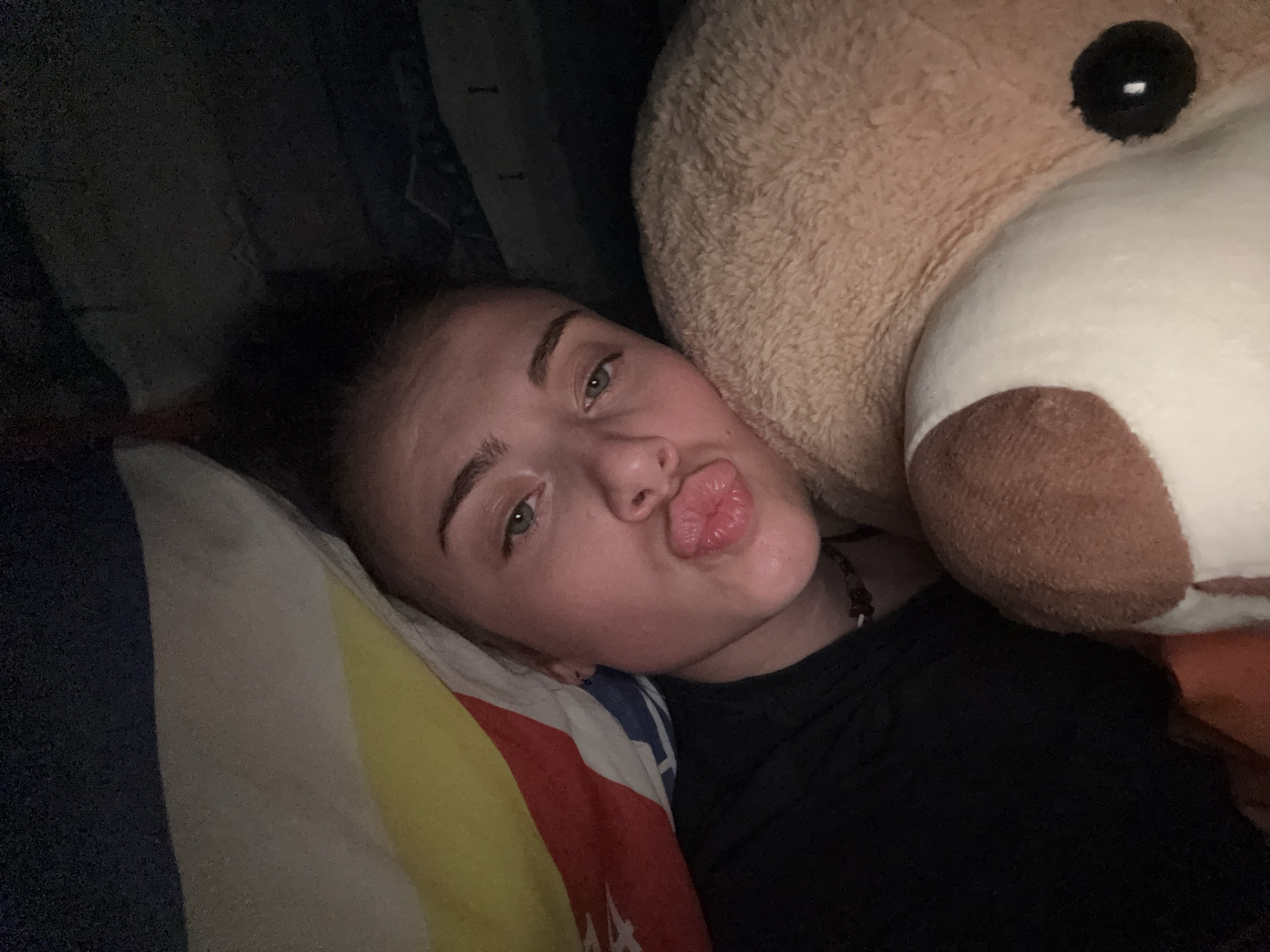 The image depicts an individual lying in a dimly lit indoor setting, positioned beside a large plush teddy bear. The person, appearing to be a young female, has fair skin, light-colored eyes, and dark hair, and is wearing a dark, possibly black, top. Her head rests on a multi-striped pillow featuring segments of dark blue, light gray/off-white, yellow, and red. Her lips are pursed in a "kissing" gesture, and her gaze is directed towards the viewer. A small earring is visible in her left ear, and a beaded necklace is partially discernible around her neck. The large teddy bear, light brown in color with a white muzzle and a black, round eye featuring a white reflection, occupies the right side of the frame. The background is dark and indistinct, suggesting low ambient light. There are no discernible elements within the image that indicate a specific geographic location, such as the city of Sofia, Bulgaria.