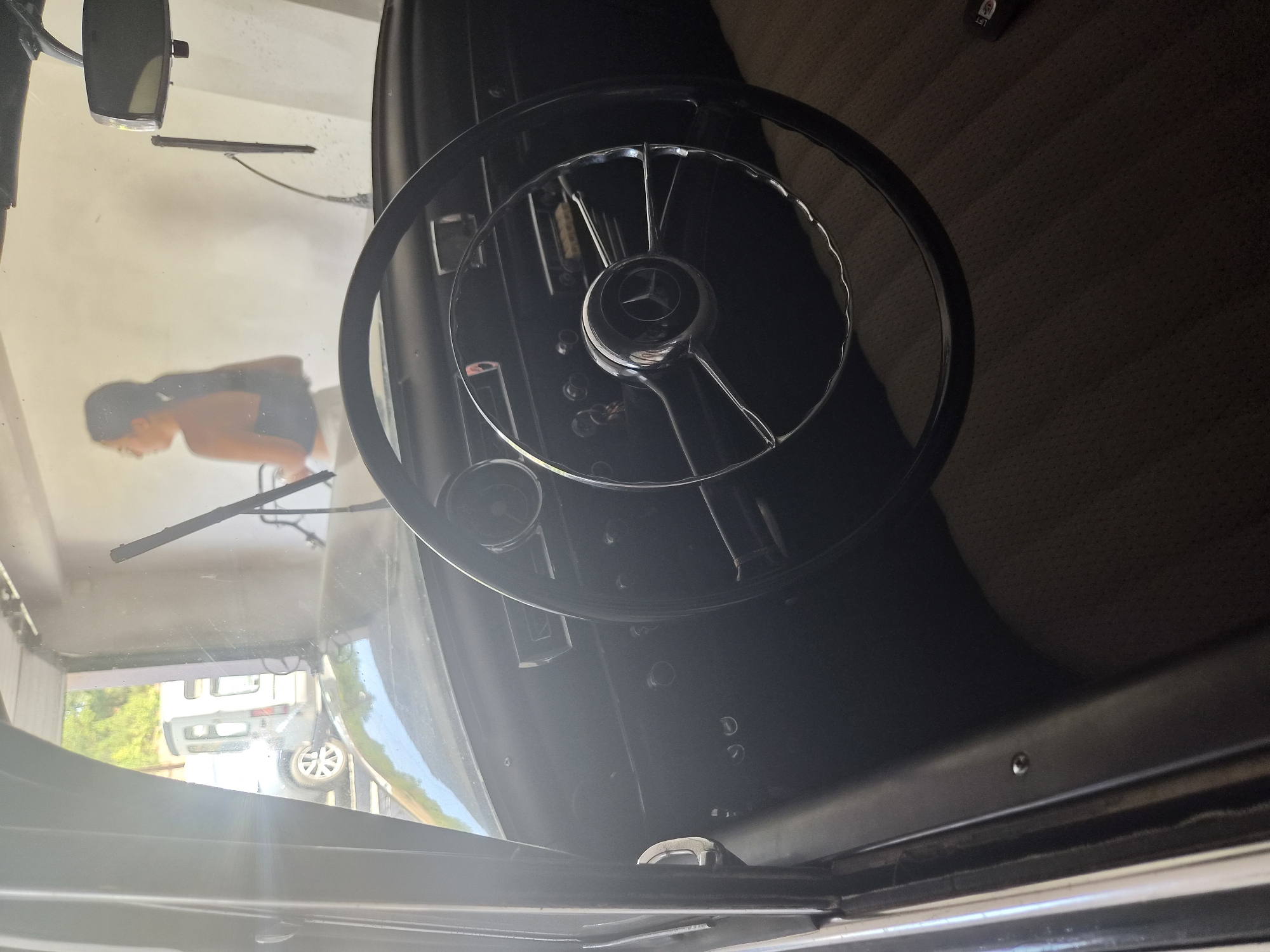 An interior shot from the driver's seat of a vintage car, possibly a Mercedes-Benz, reveals a dashboard with a classic steering wheel and chrome accents. Through the windshield, a woman is visible in the background, appearing to be washing a car in what looks like a self-service car wash. The environment suggests an outdoor or semi-covered space, with natural light filtering in. The car is parked inside a structure, and a white van is visible through an open doorway. The scene implies a mundane activity of car maintenance.