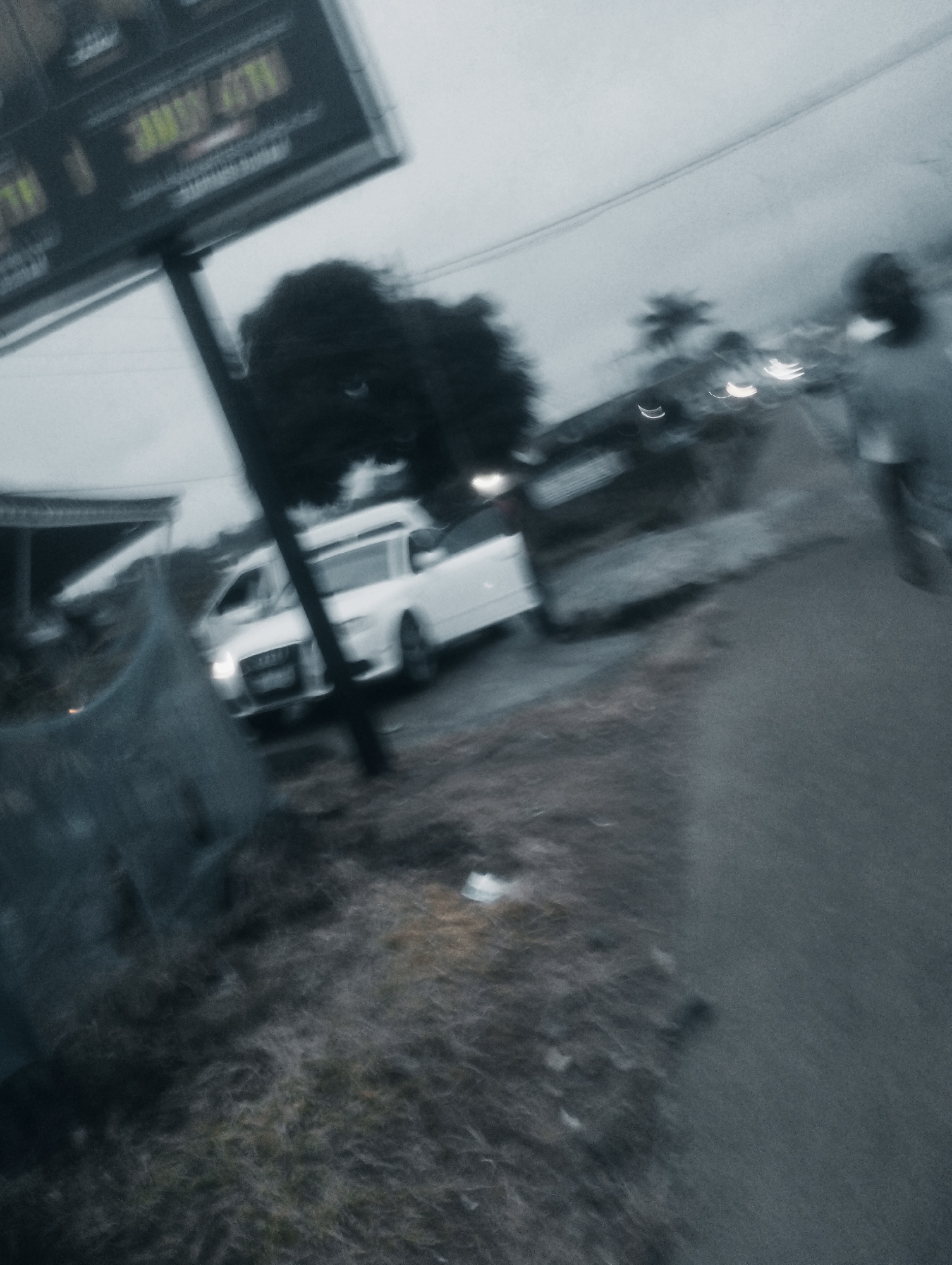 This is a motion-blurred photograph taken outdoors in what appears to be a semi-urban or roadside setting in Jos, Nigeria. The image is dominated by a grey, overcast sky and a dark, wet-looking road or path.
In the foreground, there is a blurry patch of dry grass or scrub. To the left, a large, dark, billboard-like structure is visible, angled upwards and to the left. The text on the billboard is illegible due to the blur and the image's overall low resolution. A vertical pole, possibly supporting the billboard, cuts through the frame.
Further into the scene, a white vehicle, possibly an Audi, is parked or moving slowly. Its headlights are illuminated. To the right of the car, a person is walking away from the camera, appearing as a dark silhouette. Their form is indistinct due to motion blur.
In the background, trees and other blurry shapes suggest buildings and more vegetation. The overall impression is of a slightly gloomy, possibly damp, day, with limited visibility due to motion blur and atmospheric conditions. The scene appears to be on the side of a road, with a pedestrian walkway or shoulder visible.