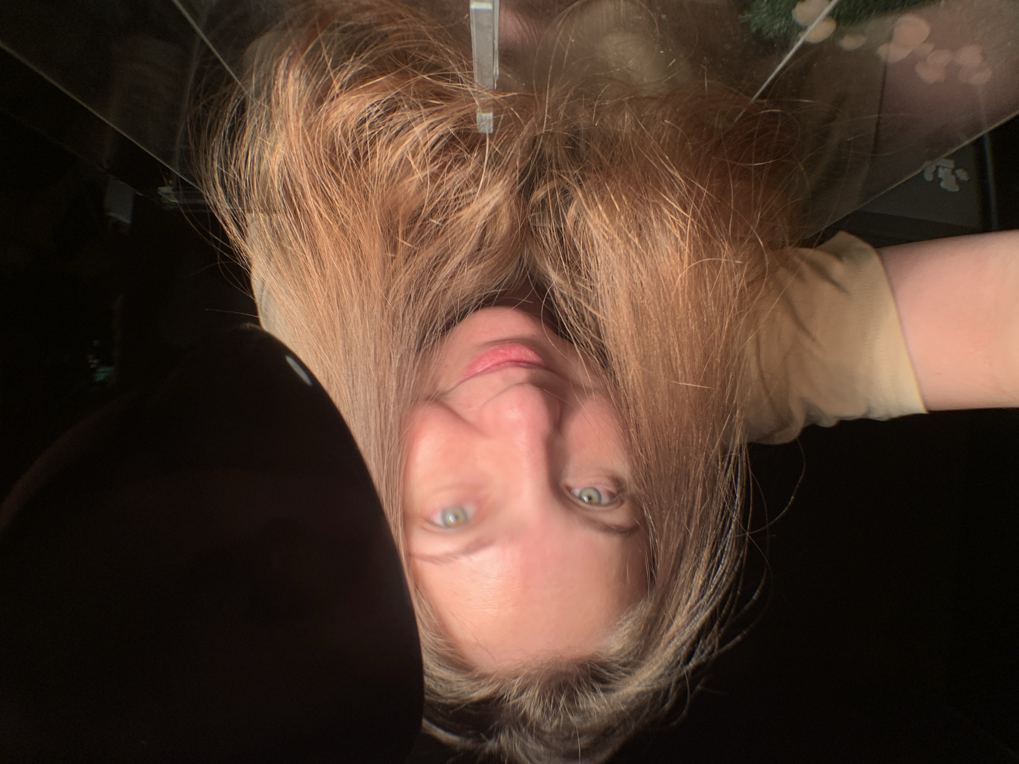 A selfie, taken upside down, shows a Caucasian woman with long, blonde hair. Her face is fair-skinned with light green eyes. She appears to be lying down, looking up at the camera, which is positioned above her. A section of her upper body, wearing a light yellow shirt, is visible in the upper right corner. A black, undefined object is located in the lower left corner. Overhead, there's a light fixture or some type of reflective surface above her. The image does not contain enough data to specify its location in the city of Chaikovsky, Russia.