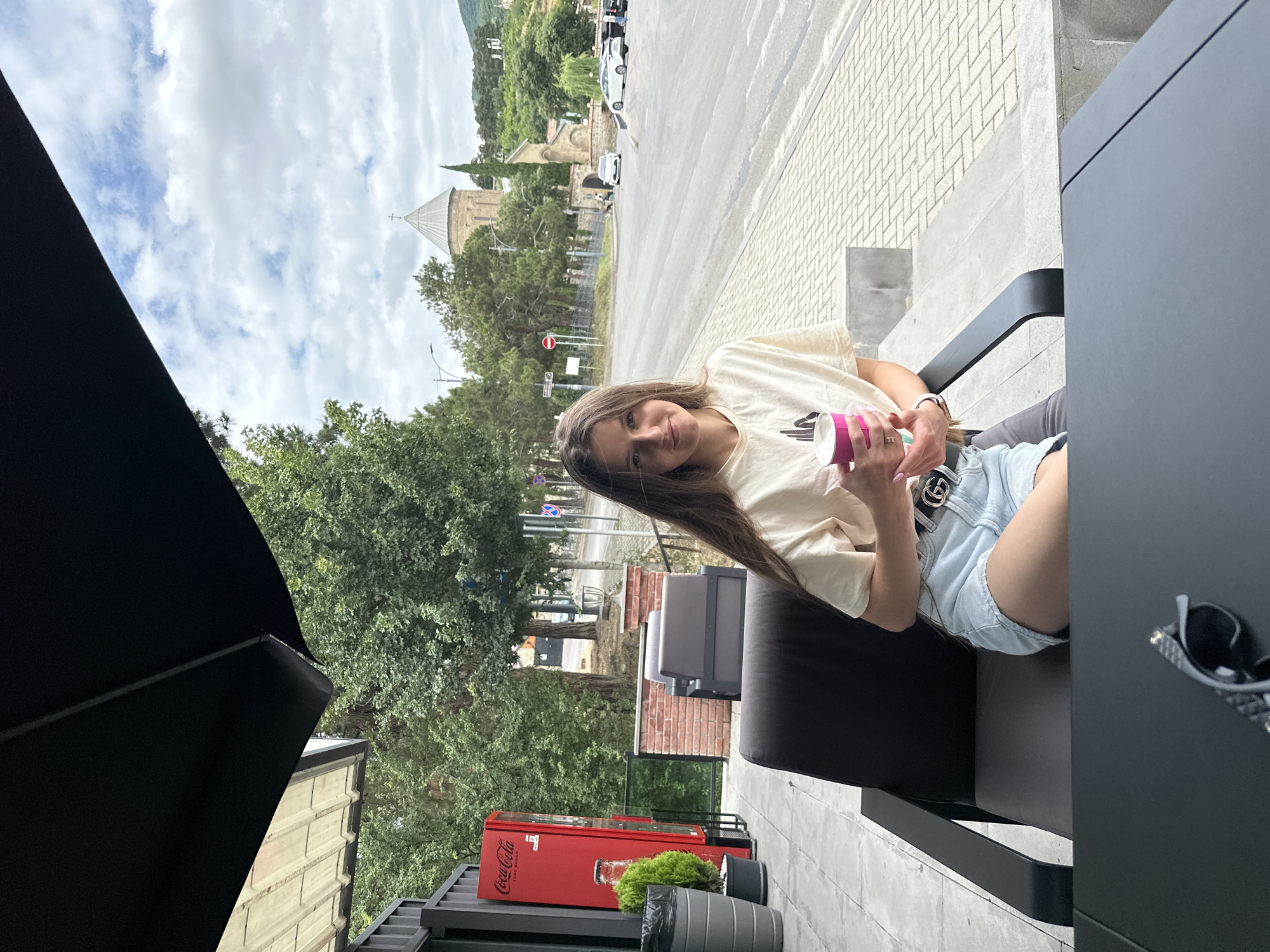 A young woman with long brown hair sits on an outdoor chair, holding a pink paper cup. She wears a light-colored t-shirt and denim shorts. Behind her, a red Coca-Cola vending machine stands next to some green bushes and a brick wall. Further in the background, a prominent stone church with a domed roof, likely part of the Svetitskhoveli Cathedral complex, is visible on a hill. The scene is set on a cloudy day, with trees and a road with cars in the mid-ground. A black table with sunglasses on it is in the foreground. The setting appears to be an outdoor seating area of a cafe or restaurant in Mtskheta, Georgia.
