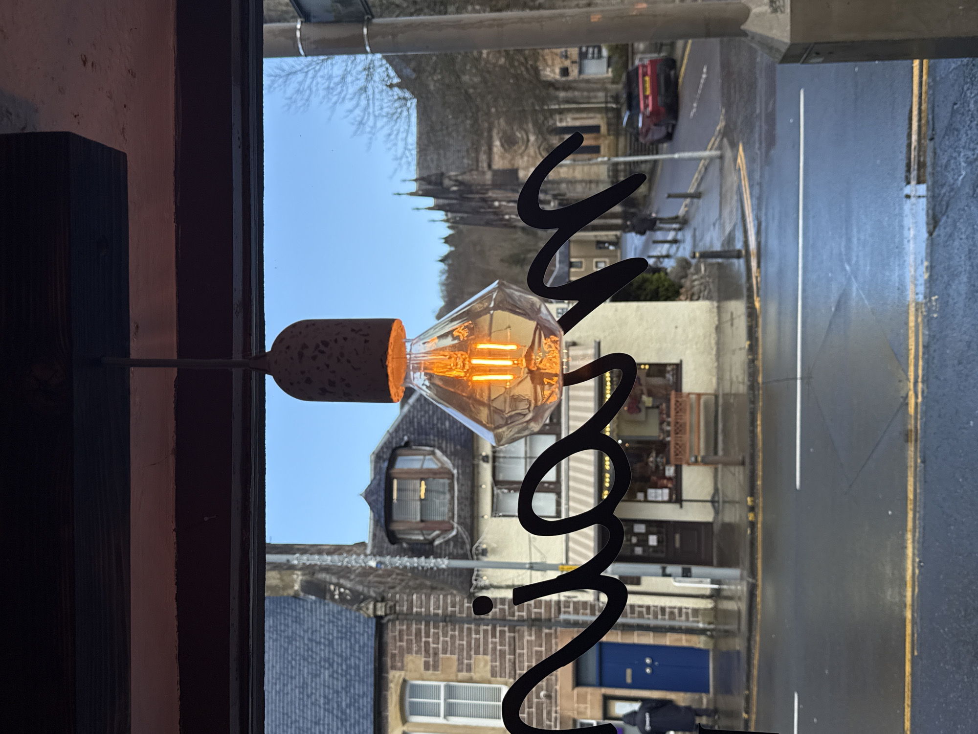 The image displays an exterior urban scene viewed through a window, featuring an illuminated light fixture and partial lettering on the glass in the foreground.
In the immediate foreground, a geometric, faceted light bulb with an amber glow hangs from a rustic-textured concrete-like base, suspended from a dark wooden beam. Below the beam, black cursive lettering is affixed to the window, spelling out segments such as "...nia M".
Beyond the window, a wet, dark asphalt street with visible white lane markings and double yellow no-parking lines curves gently to the right. Wet sidewalks flank both sides of the street.
Along the street, various buildings are present:
To the left, a two-story structure constructed of light brown brick features a prominent blue door and white-framed windows.
In the center, a cream-colored rendered building houses a ground-floor shop with a dark brown wooden door, a striped awning, and a wooden bench placed outside. Above this, a distinctive bay window with multiple vertical panes projects from the second story, topped with a dark, conical roof.
Further down the street and to the right, a large, dark stone church with multiple spires, exhibiting Gothic architectural characteristics, stands against the horizon.
Several individuals are present on the street: one person in a dark hooded coat stands near the blue door on the left, facing away. Another person in dark clothing walks further down the street near the central building.
A red Range Rover Evoque SUV is parked or moving slowly down the street to the right, facing away from the viewer.
Dark bollards line the sidewalk, and a grey utility pole is visible on the right side of the frame.
In the background, bare deciduous trees cover distant hills under a clear blue sky.
The location is a city street in Unknown, Unknown, under ambient daylight conditions following recent precipitation.