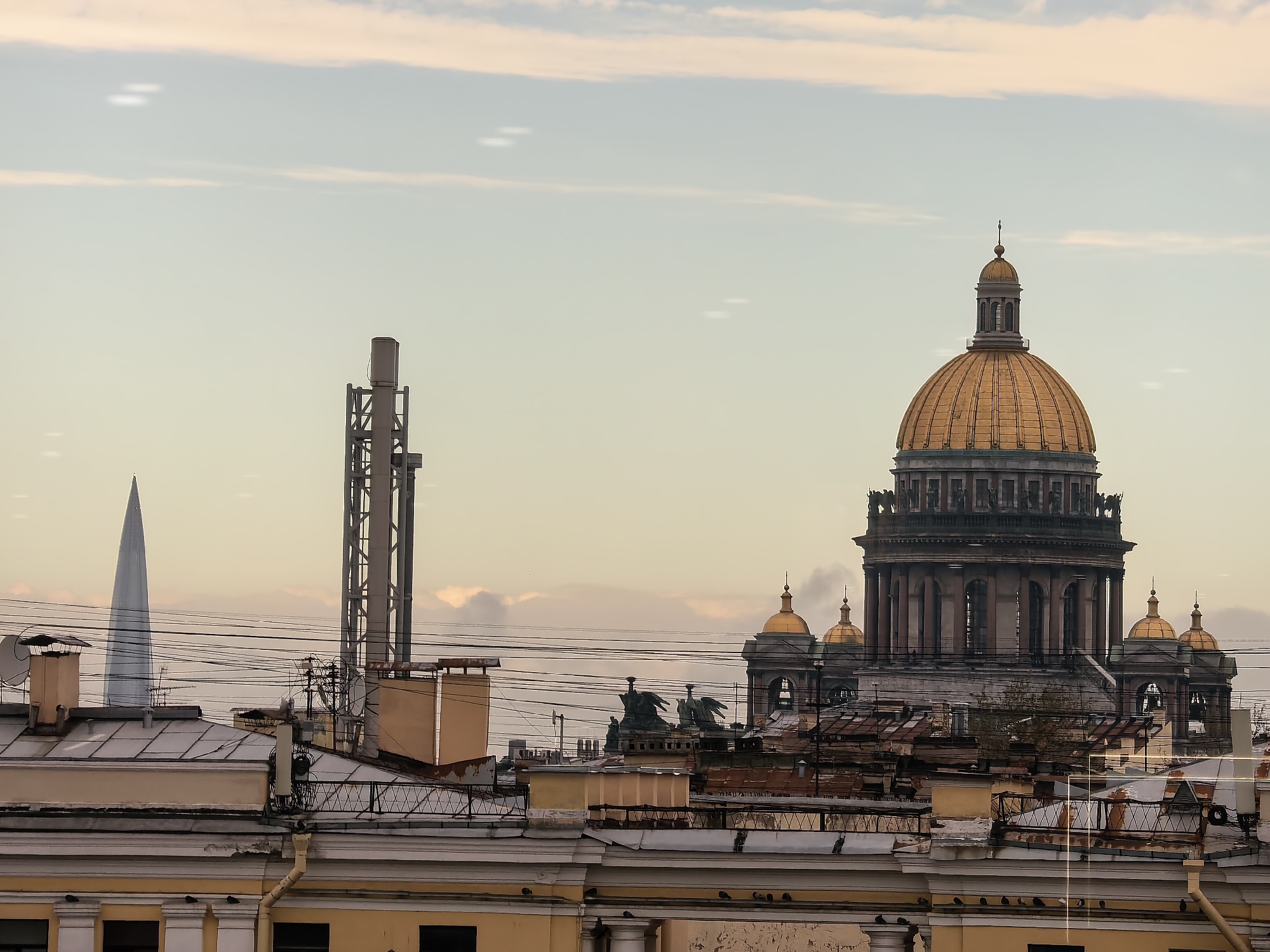 The image captures a cityscape in Saint Petersburg, Russia, likely during the late afternoon or early evening, given the soft, warm light. The most prominent structure is the golden dome of Saint Isaac's Cathedral, a grand landmark of the city, which dominates the right side of the frame. In front of the cathedral, a dense collection of rooftops with various architectural features, including chimneys and satellite dishes, creates a layered foreground. Several power lines crisscross the sky above these buildings.
To the left of the cathedral, a tall, modern glass skyscraper, resembling the Lakhta Center, pierces the sky. A large, industrial-looking metal structure, possibly a communication tower, stands between the skyscraper and the cathedral. The sky above is a pale, hazy blue with streaks of pink and orange, indicative of sunset or sunrise. There are a few blurry white spots in the sky, which could be lens flares or atmospheric anomalies. No people are visible in the image. The overall impression is one of a city where historical architecture and modern infrastructure coexist.