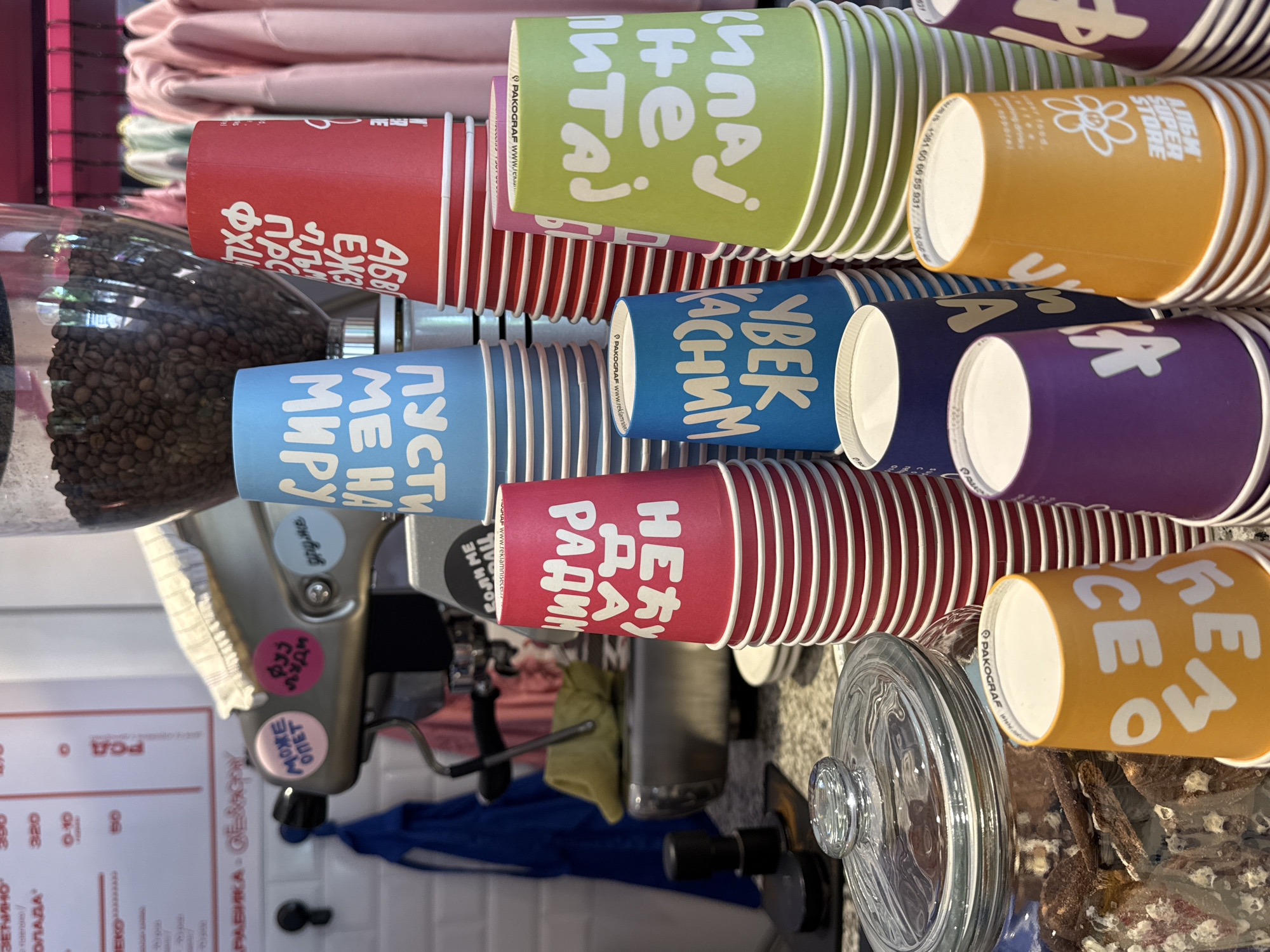 The image shows a stack of colorful paper coffee cups in what appears to be a coffee shop or cafe. The cups are stacked in multiple rows and have different colors, including red, green, blue, purple, and orange. Some of the cups have writing in Cyrillic, suggesting the location is in a Slavic country. There is a coffee grinder with beans visible, along with a coffee machine. A menu board in the background lists drink prices. An orange cup has the name "Albi Super Store". The word "Ravnica-Selo" is also printed, suggesting this location is a part of Novi Sad, Serbia.