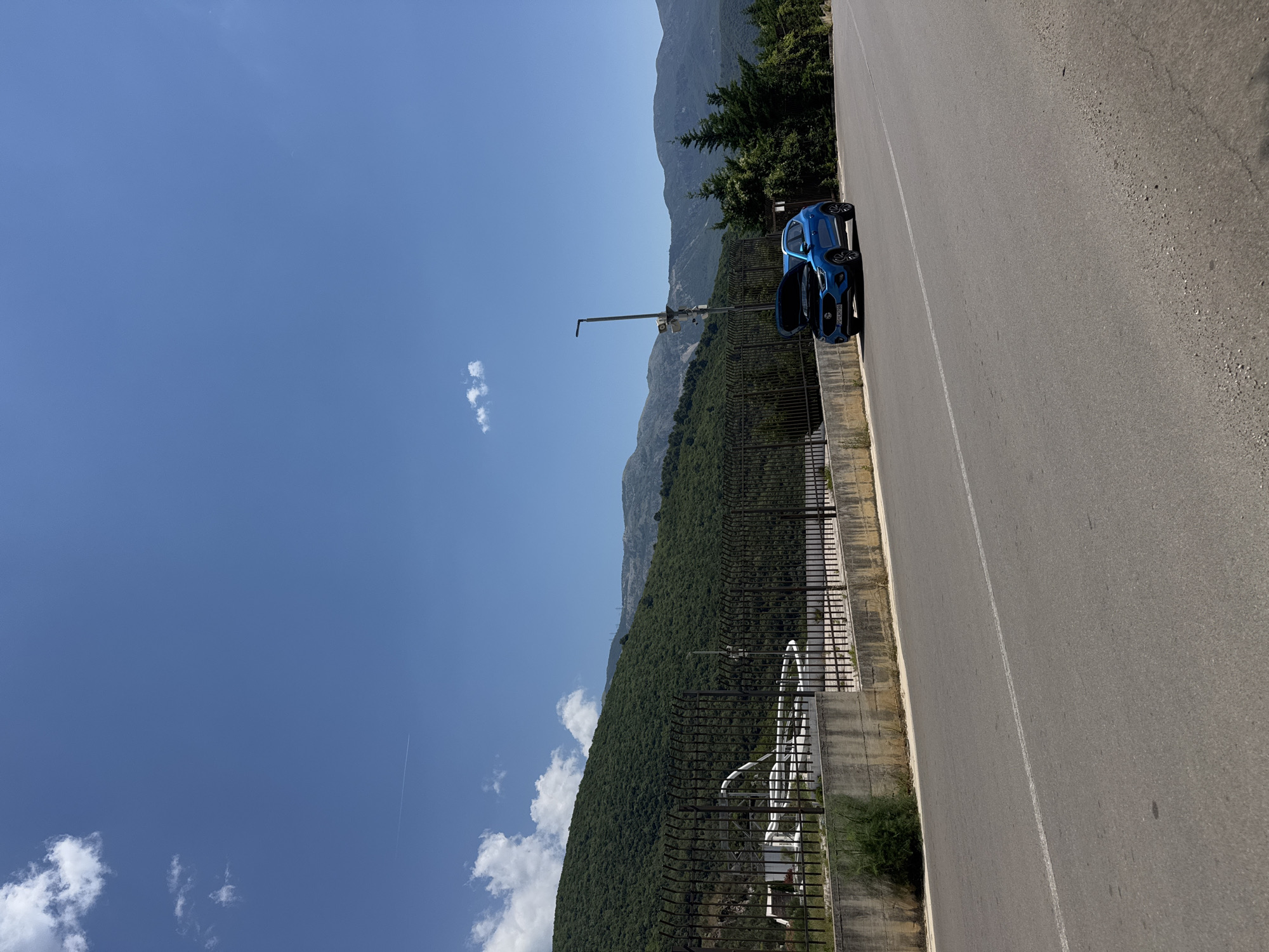 A blue car with its hood open is parked on the side of a paved road in a mountainous, rural area. The car is positioned to the right of the frame, on the shoulder of the road. Behind the car, a dark metal fence runs horizontally across the image, separating the road from a green, tree-covered hillside. Beyond the hill, more mountains are visible in the distance under a clear blue sky with scattered white clouds. A single white contrail is visible in the upper left quadrant of the sky. A lamppost with attached cameras stands near the car, just behind the fence. The scene appears to be during daylight hours.