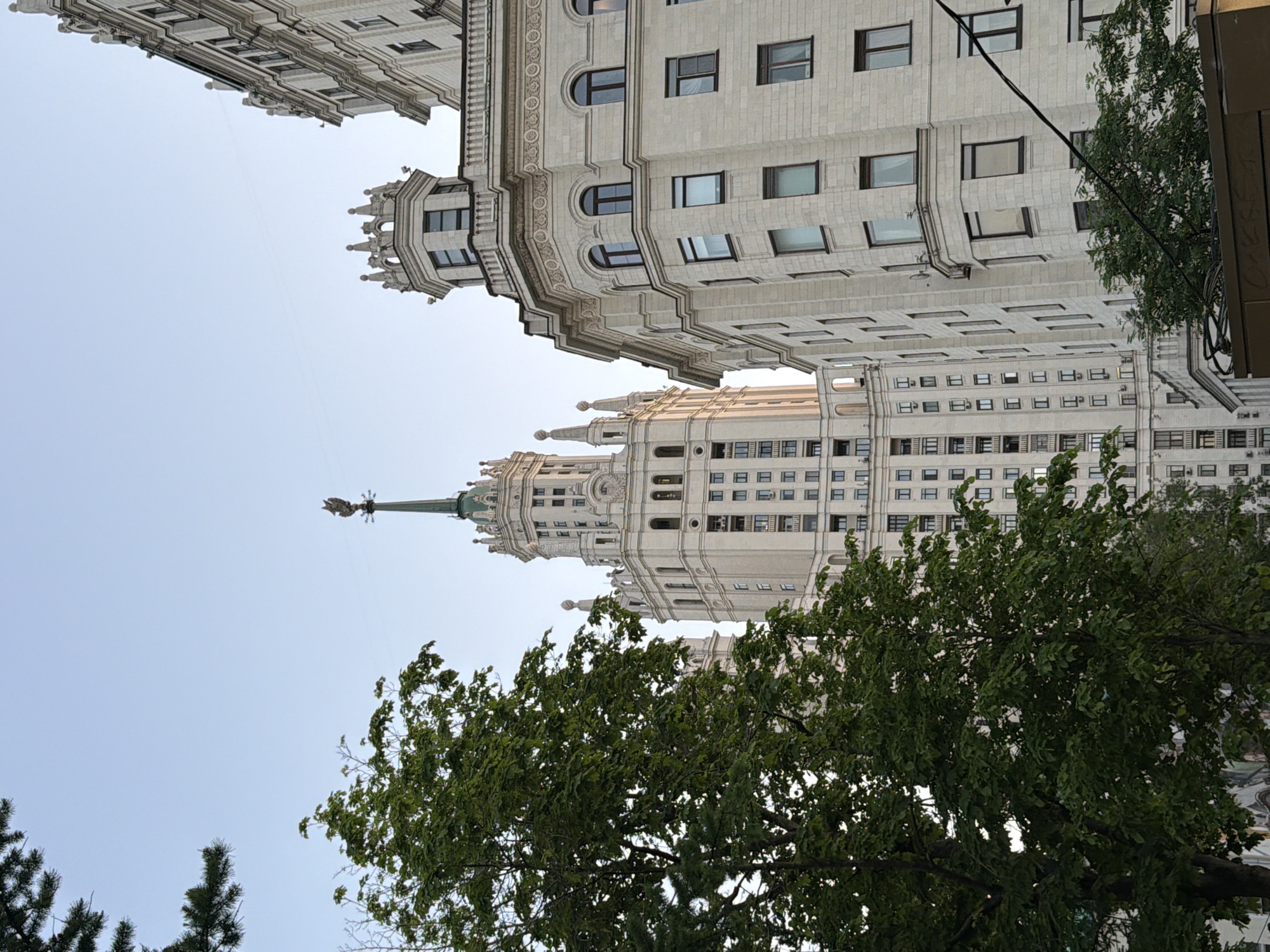 The image depicts a low-angle shot of the Kotelnicheskaya Embankment Building, one of the Seven Sisters skyscrapers in Moscow, Russia. The building has a cream-colored facade with numerous windows and decorative elements. Parts of other buildings are visible above. Trees with green foliage obscure parts of the building. The sky is overcast.