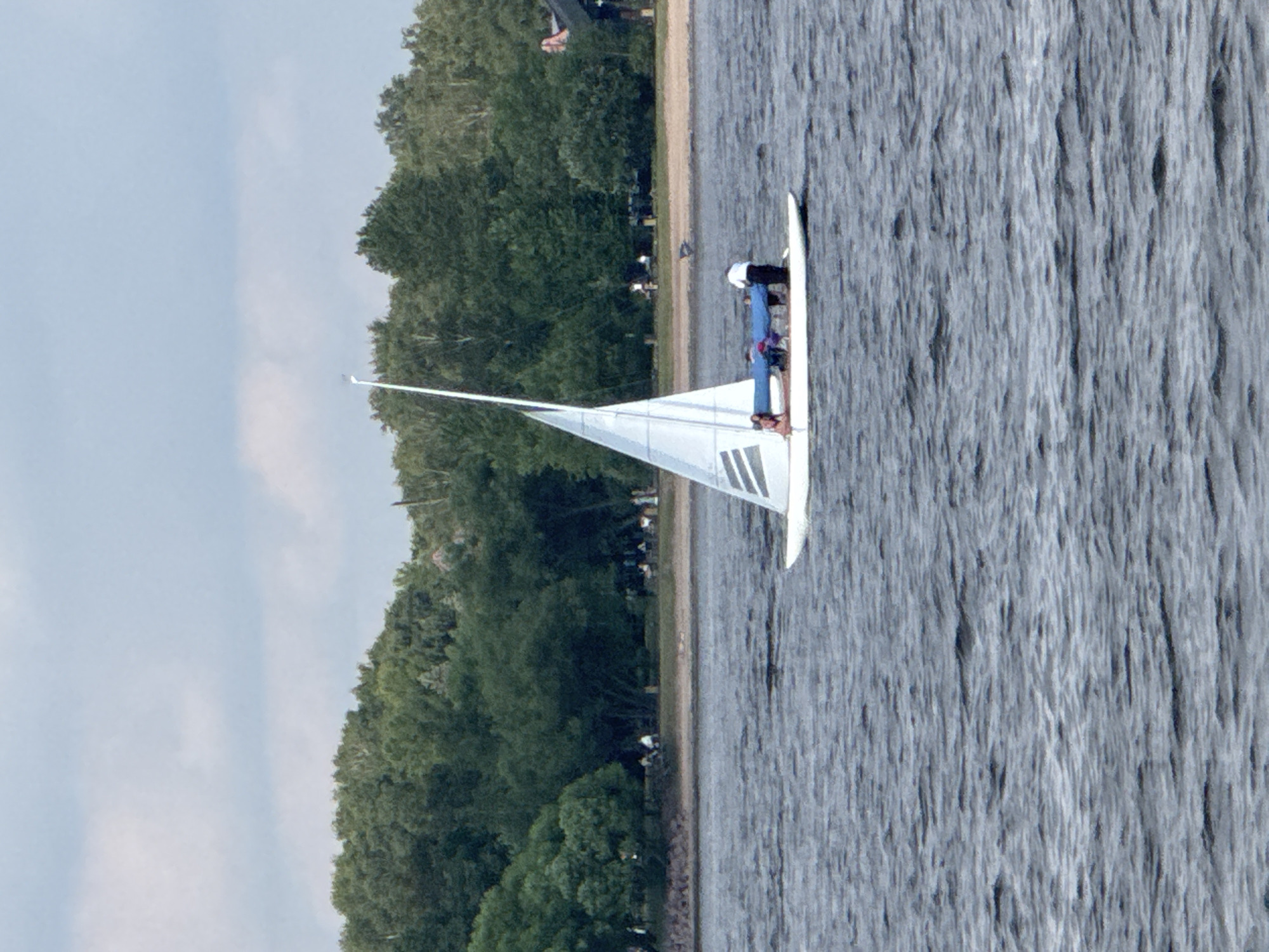 The image shows a white sailboat with a single mast and sail on a body of water. There are several people on the sailboat. In the background, there is a shoreline with trees and buildings. The sky is cloudy. The scene is likely a recreational area in Saint Petersburg, Russia, possibly on one of its many rivers or canals, given the presence of sailboats and waterfront greenery common in the city.