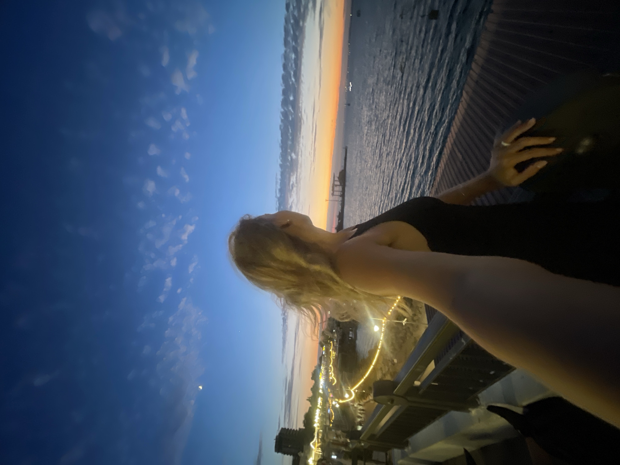 A wide-angle, low-light photograph captures a young woman with blonde hair, seen from the chest up, looking out towards a body of water and a city skyline at dusk. She is wearing a black tank top and her left hand rests on a dark railing in the foreground, adorned with a ring on her left ring finger. Her right arm is extended, creating a selfie-like perspective.
The background depicts a twilight scene over a body of water, with the sun having recently set, casting a warm orange and pink glow on the horizon. The sky above is a deep blue, transitioning to lighter shades near the horizon, with scattered wispy clouds. A crescent moon is visible in the upper left quadrant of the sky.
To the left of the frame, a waterfront promenade is illuminated by strings of golden lights, suggesting a lively evening atmosphere. Buildings are visible along the shoreline, including a darker, taller structure in the far distance. The water itself is calm, with subtle ripples reflecting the ambient light. The location appears to be a coastal area, potentially a pier or promenade, with a dark metal railing in the foreground and a wooden deck with horizontal planks beneath the woman's hand and the railing.
The overall impression is one of a serene evening in a coastal city, with the woman enjoying the view. Given the context provided, this scene is likely located in Anapa, Russia, a popular resort town on the Black Sea coast, known for its beaches and promenade.