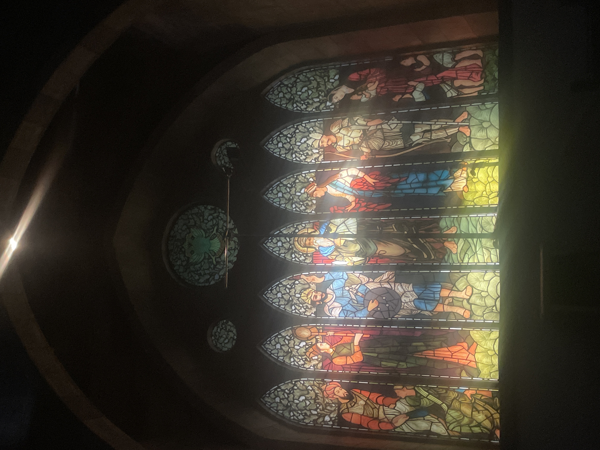 The image shows a tall, arched stained glass window with multiple sections depicting figures. The window is situated within a dark, possibly interior, space with stone or brick walls. The stained glass panels are colorful and appear to depict scenes with people in historical or religious attire. There are circular glass ornaments to the left of the stained glass panel. The image is taken from an angle that makes the stained glass window the prominent subject. The location is unknown and not readily evident from the image.