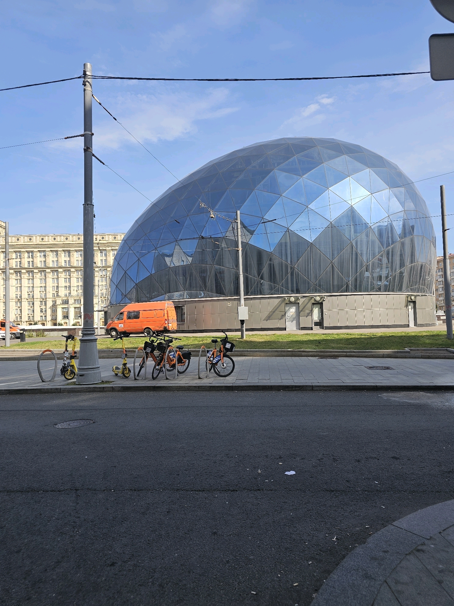 The image shows an outdoor scene with a large, modern building with a glass, geodesic dome-like facade. In front of the building, there is a row of parked rental bikes and scooters. An orange van is parked in front of the building, and a tall utility pole with wires stands in the foreground. In the background, there is a large, light-colored apartment building. The scene appears to be in a city, likely in the morning or afternoon, given the sunlight and clear blue sky with a few clouds. The ground is paved with asphalt and concrete. This location is in the city of Khimki, Russia, specifically at the Arkangel stadium.