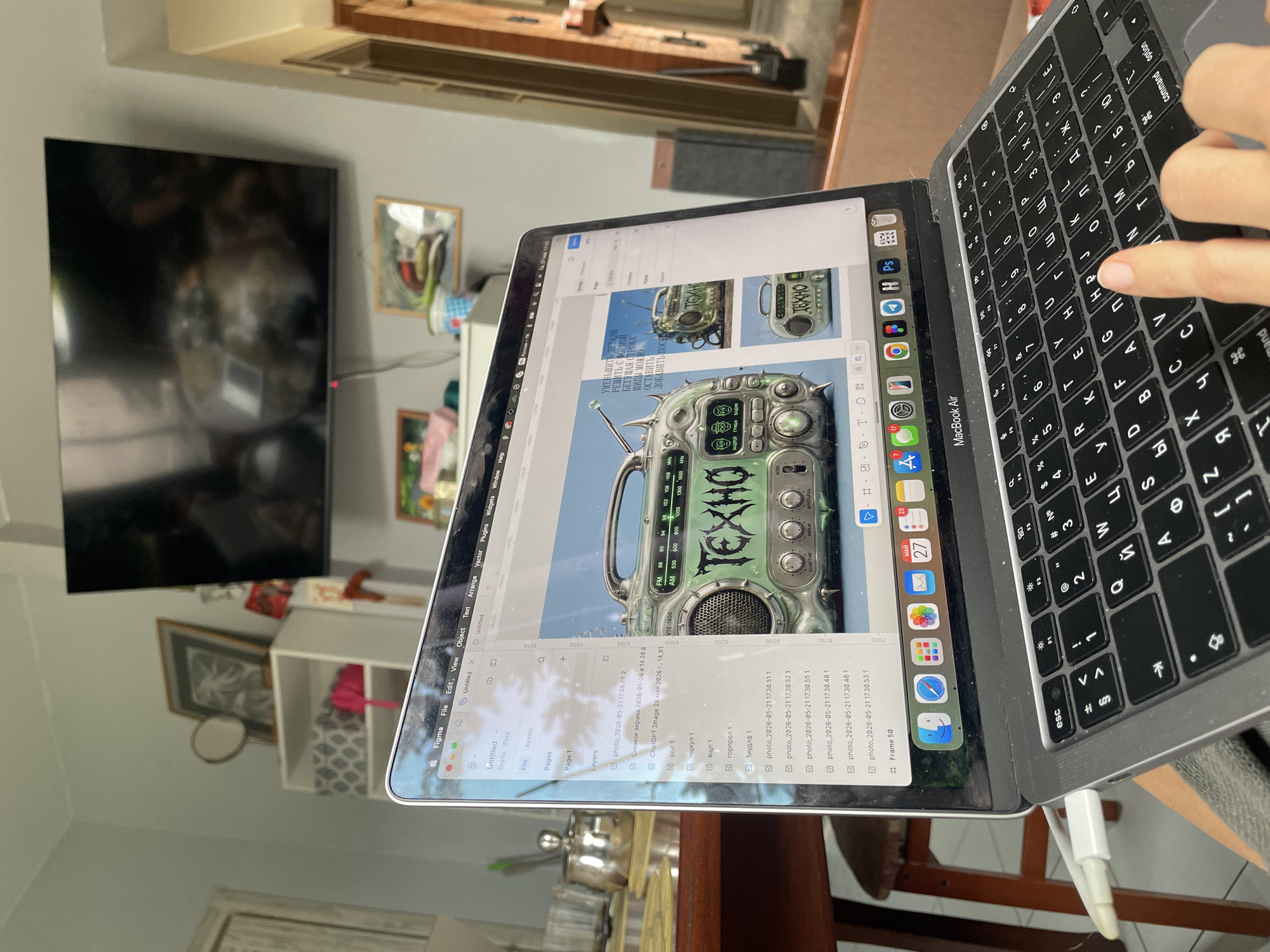 A person is seated at a wooden table, typing on the keyboard of a Macbook Air laptop. The laptop screen displays the Figma design software, showcasing a graphic design project that features an illustration of an antique radio with the word "TEXHO" written across its front. The keyboard layout is Cyrillic. The laptop is connected to a white charging cable.
In the background, a television is mounted on the wall. A shelving unit with various items, including a picture frame and a pink object, is visible below the television. The table is partially cluttered with miscellaneous objects. The laptop is located in a building with a wooden door frame, possibly in a home or office setting. The location in the city of Sestroretsk, Russia is not discernible from the image.