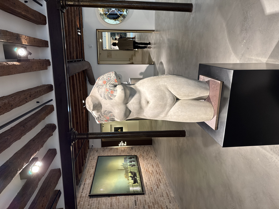 This image depicts the interior of an art gallery or exhibition space located in Venice, Italy, characterized by a blend of historic and modern architectural elements.
In the foreground and center, a full-length, headless, and legless stone sculpture of a female torso is displayed. The sculpture is a pale grey, possibly concrete or light stone, and features detailed, dark grey tattoo-like patterns on its shoulders and upper arms, depicting stylized waves or clouds and clusters of small red flowers. It rests on a black rectangular plinth, which is topped with a thin, reddish-brown square base.
To the left, a large, rectangular framed painting hangs on a exposed brick wall. The artwork depicts a tranquil Venetian canal scene with several gondolas and historic buildings silhouetted against a luminous, hazy, pale yellow and green sky.
In the midground to the right, an open doorway reveals a person walking away from the viewer. This individual, appearing male, wears dark trousers and a light brown or khaki jacket. Through the doorway, part of another artwork, possibly a large portrait or abstract piece, is visible. Further to the right, on a white wall, a circular, ornate art piece or decorative mirror with geometric patterns is mounted.
The ceiling features exposed, dark wooden beams and a modern track lighting system with multiple spot lights. Two dark, fluted metal columns support a thick, dark structural beam running across the ceiling. The floor throughout the space is a smooth, polished grey concrete. The overall environment is well-lit and appears to be a contemporary exhibition setting within an older building.