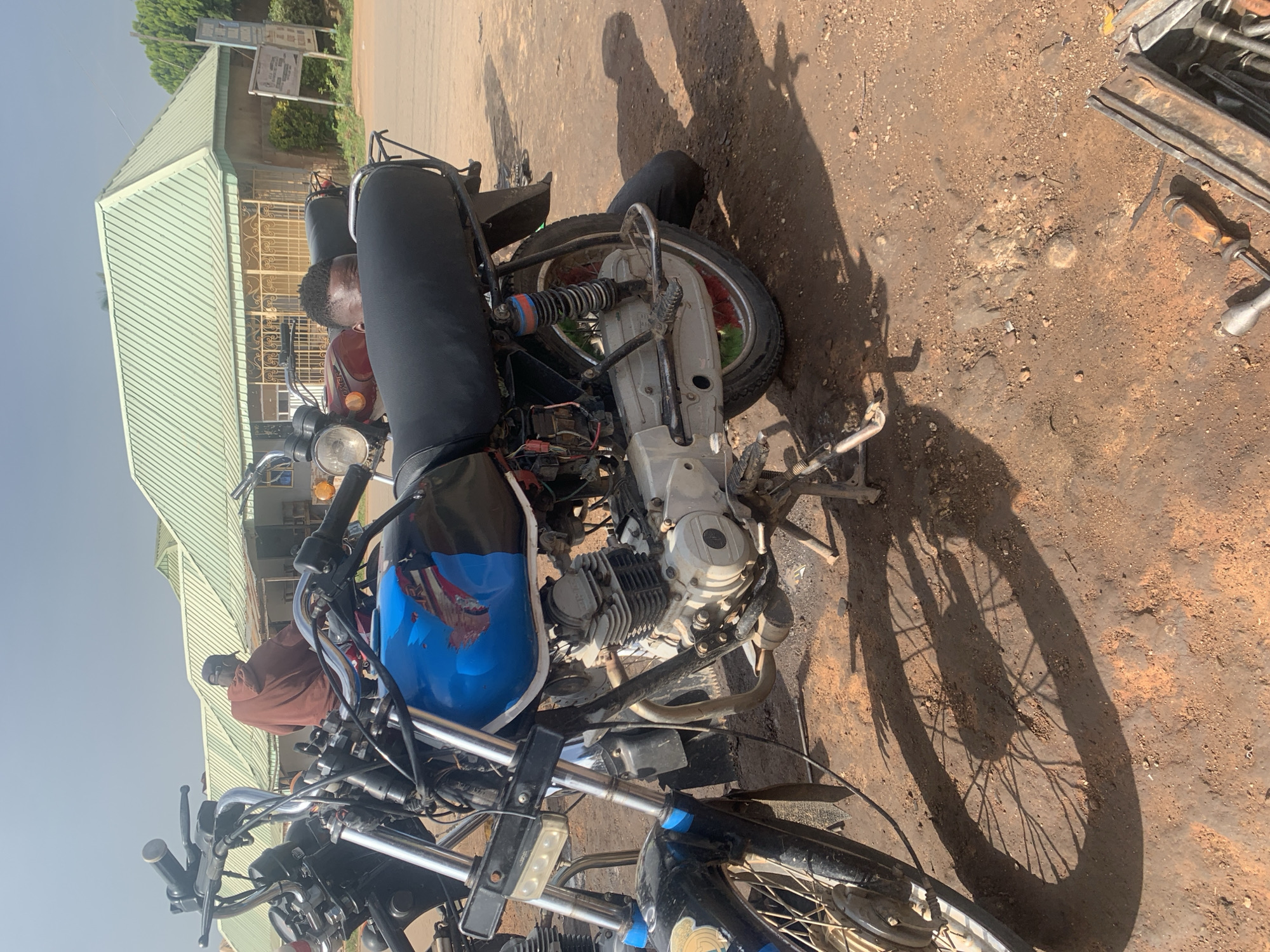 The image depicts a dusty outdoor area in Gura Top, Nigeria, likely a mechanic's workshop. Several motorcycles are present. In the foreground, a blue and black motorcycle is prominently displayed, its engine and wheels clearly visible. A person is partially obscured behind the seat of a red motorcycle in the middle ground. Further in the background, a building with a distinctive light green, corrugated metal roof stands under a bright, overcast sky. The ground is covered in reddish-brown dirt and scattered debris, indicative of a work environment. Tools, including a socket wrench, are visible in the lower right corner. The overall scene suggests a casual, functional setting for motorcycle repair and maintenance.