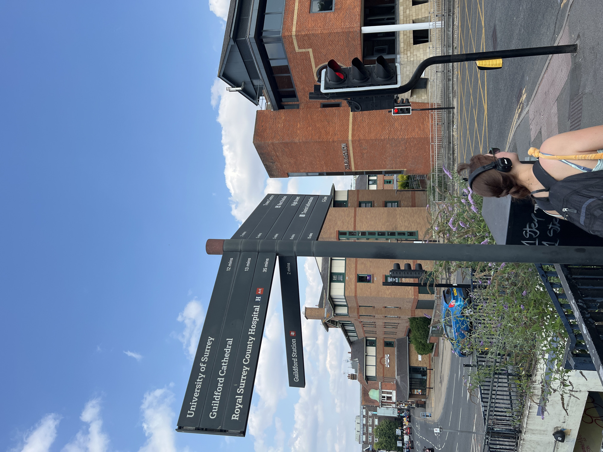 A woman wearing headphones and a backpack stands on a sidewalk in Guildford, United Kingdom, facing away from the camera and towards a street with a traffic light. In the background, there is a multi-story brick building, a blue car, and a directional signpost with text indicating "University of Surrey", "Guildford Cathedral", "Royal Surrey County Hospital", and "Guildford Station". The sky is blue with scattered clouds.