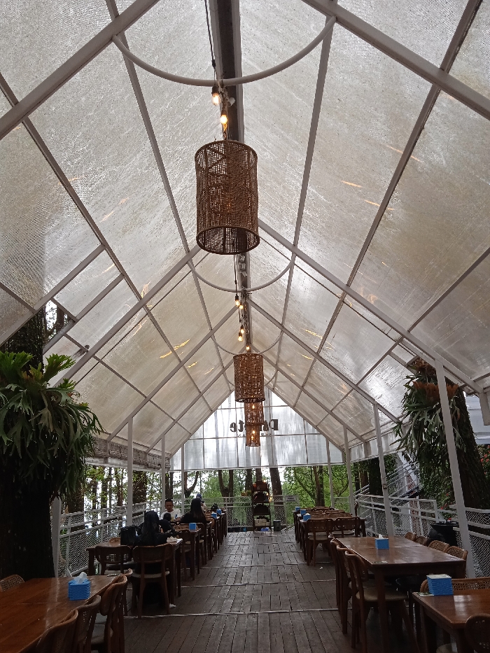 The image shows the interior of a restaurant, with a transparent roof and wooden floor. The restaurant is situated in a garden setting. The roof is made of a transparent material, possibly polycarbonate, and is supported by white metal beams, creating a grid pattern. There are hanging light fixtures with woven lampshades, lit by incandescent bulbs. The restaurant has wooden tables and chairs and several patrons are seated at tables. The chairs are mostly brown wooden chairs, with some upholstered chairs. There is a counter near the entrance with a black sign. The background shows trees and greenery, with some areas visible through the transparent roof. The restaurant is located at coordinates -7.669, 111.217, which is in Plaosan, Indonesia. The image is well-lit and the scene is captured from an angle looking towards the entrance of the restaurant.