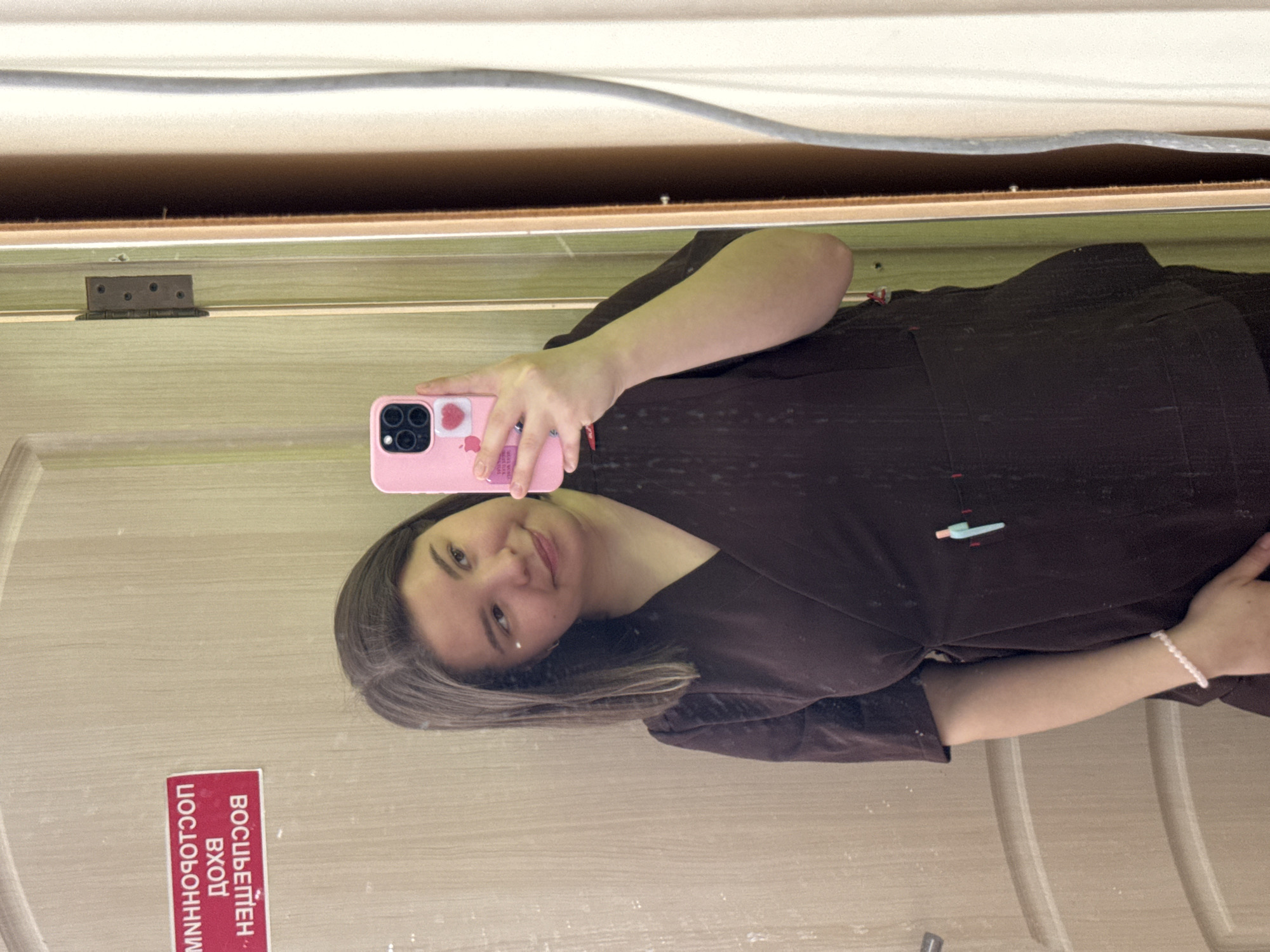 A woman is taking a selfie in what appears to be a mirror reflecting the interior of a room. She is holding a pink smartphone. She is wearing a dark brown scrub uniform. On the door frame, a sign in Russian reads "ВОСЬМЁРКИ - ВХОД ЧОСОБОНННМ". The presence of the Russian text suggests the location is likely in Russia. Based on the sign, the location is possibly in Kazan, Russia, due to the unique linguistic spelling of the text "ЧОСОБОНННМ". The environment is relatively plain and unremarkable, suggesting a functional space.