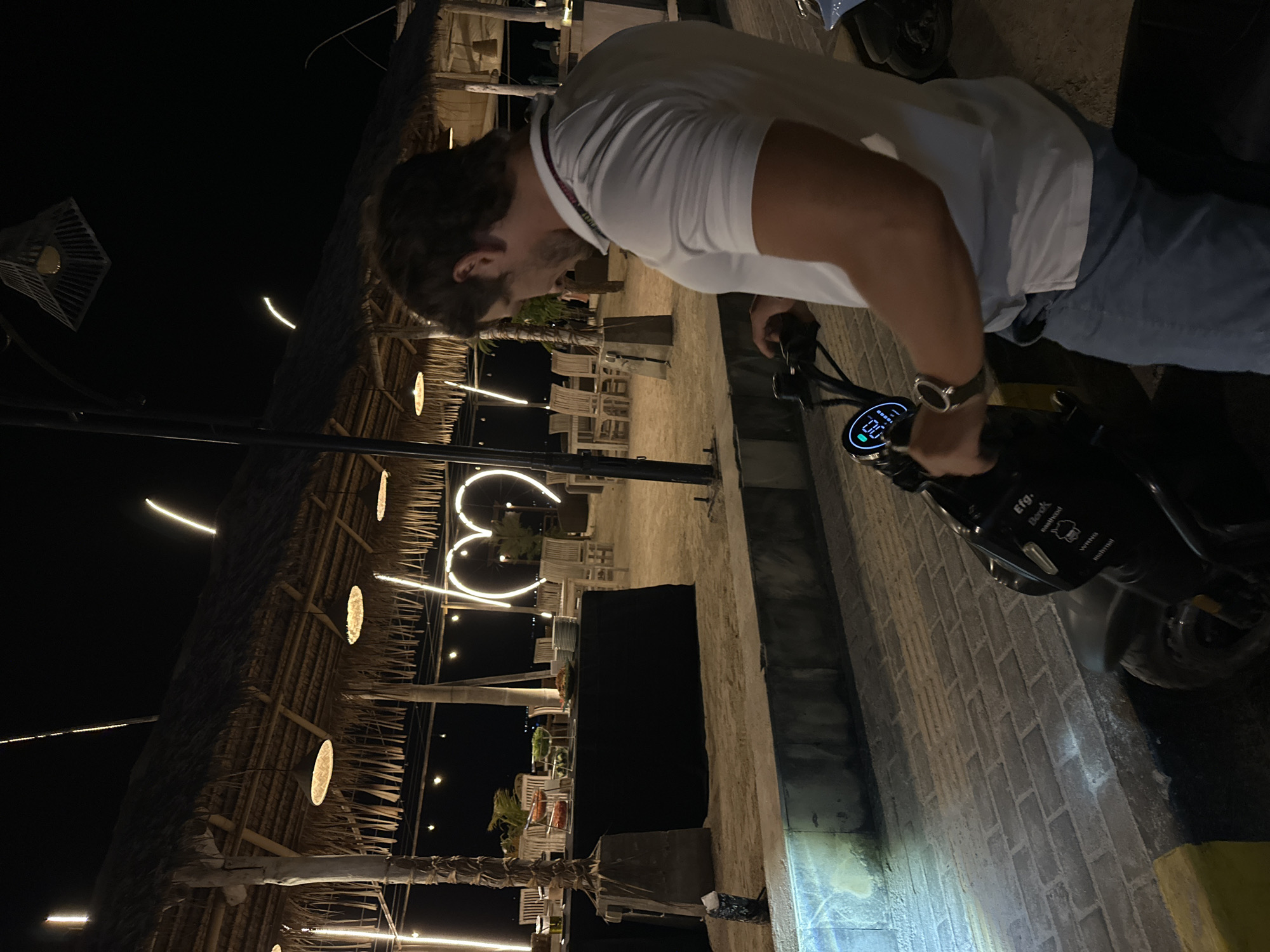 A person is standing next to a motorized scooter at night in what appears to be an outdoor dining area. The person is wearing a white shirt and blue jeans, and they appear to be operating the scooter's controls. The scooter has a visible digital display. The dining area has a thatched roof, string lights, and heart-shaped lighting decorations. The location is the city of Unknown, Indonesia.