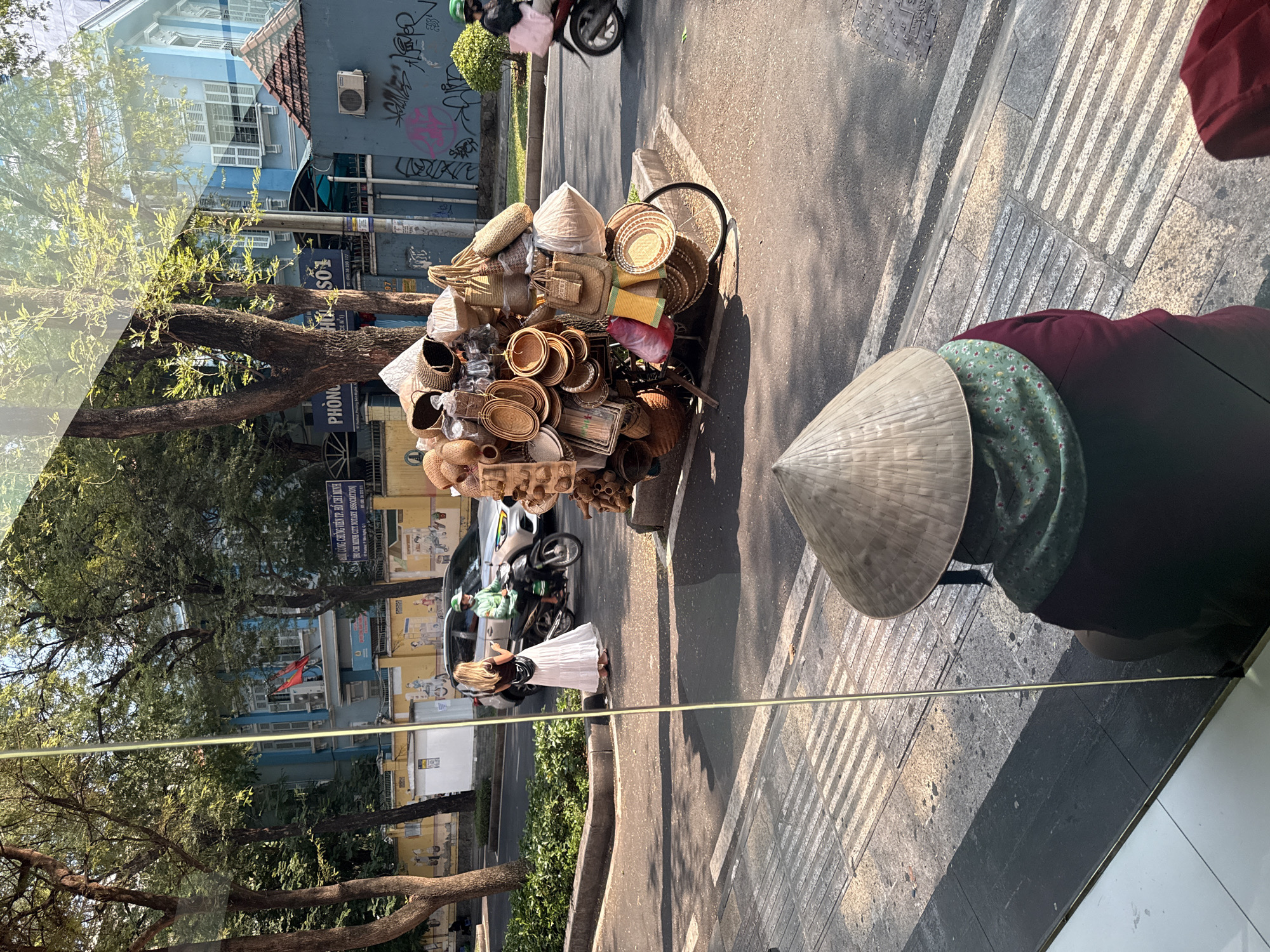 The image shows a street scene in Thủ Đức, Vietnam, with a woman wearing a traditional conical hat walking on a sidewalk with tactile paving. Next to her, a bicycle laden with various woven baskets and conical hats sits on the edge of the road. A person in a white dress is walking on the other side of the street. Cars and motorbikes are visible on the road. Buildings and trees line the street, and signs with Vietnamese text are visible on the buildings.