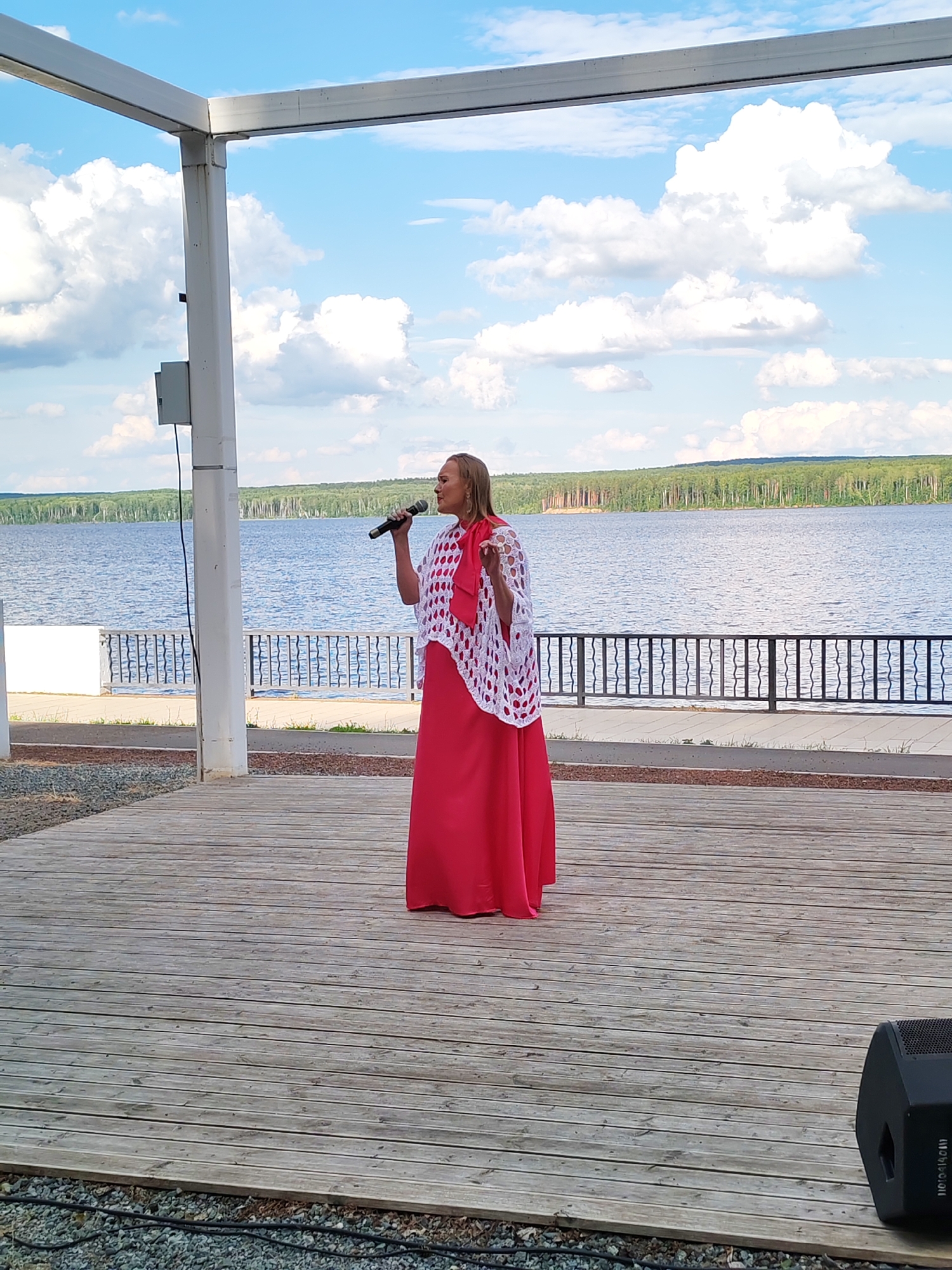A woman is singing into a microphone on a wooden outdoor stage. She is wearing a long, bright pink dress and a white crocheted shawl. Behind her, a calm body of water stretches to a tree-lined horizon under a partly cloudy blue sky. The stage is located outdoors, likely at a waterfront venue in Chaikovsky, Russia, as suggested by the environment. A black speaker is visible on the lower right side of the stage. The overall scene depicts an outdoor performance event.