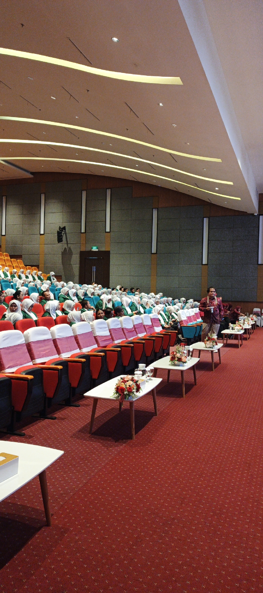 The image shows an indoor event held in a convention hall or auditorium in Surabaya, Indonesia. The room has tiered seating with red and white chair covers, occupied by a large audience, predominantly women wearing green tops and white hijabs. In the foreground, there are several white tables with flower arrangements and glasses of water. A man in a patterned shirt stands near the tables, facing the audience, appearing to be a speaker or presenter. The ceiling features modern lighting and architectural elements. The overall atmosphere suggests a formal gathering or conference.