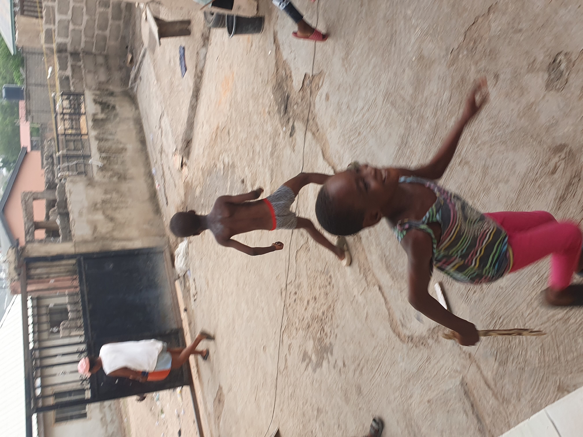 This image captures an outdoor scene in a residential or communal area, likely in Lokoja, Nigeria, under bright daylight. The ground is an uneven, light-colored concrete or dirt surface.
The primary subjects are three children and an adult, engaged in various activities. In the foreground, a young girl is seen in dynamic motion, wearing a multi-colored striped sleeveless top and pink leggings. She is smiling or laughing, suggesting joy, and holds a long, thin stick in her right hand, indicative of play. Behind her, slightly to the left, another child, a boy wearing dark shorts with a red stripe, is also in motion, appearing to run or walk across the ground. A third child's head and torso are partially visible further back on the right side of the frame.
In the middle ground, to the left, an adult or older person stands near a tall, dark metal gate, wearing a white top over dark shorts and a light head covering. The gate is made of dark metal bars and sheets, with concrete block walls of a building visible behind it. Further back, a building with a reddish-brown roof and some distant greenery are also visible. A black water tank on a metal stand is situated above the buildings on the left side. A long, dark cable or rope stretches across the ground from the middle to the lower right of the frame.
The scene conveys a sense of everyday life and children at play. The bright illumination indicates it is daytime. No visible text is present in the image.