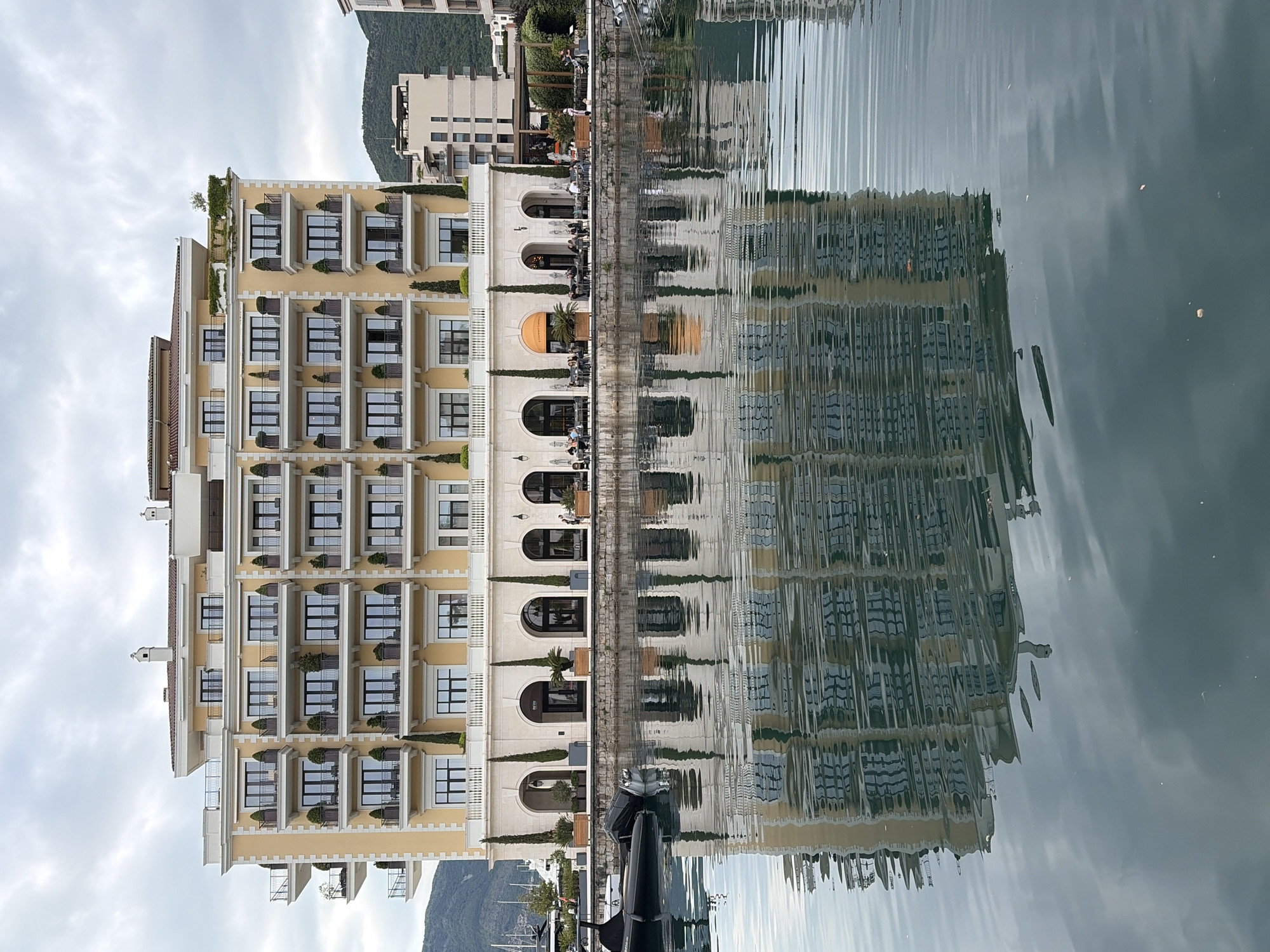The image captures a vertical view of a waterside setting in Tivat, Montenegro. A multi-story building is prominently displayed on the left, featuring a light yellow facade with rows of windows, balconies, and architectural details. Adjacent to the building is an arcade with arched openings, behind which people are seated, suggesting a restaurant or cafe.
The scene is mirrored in the water, creating a symmetrical effect. The water surface is slightly rippled, causing a distortion of the reflected image.
A mountain is visible in the background. The sky is overcast. A boat is partially visible in the foreground.