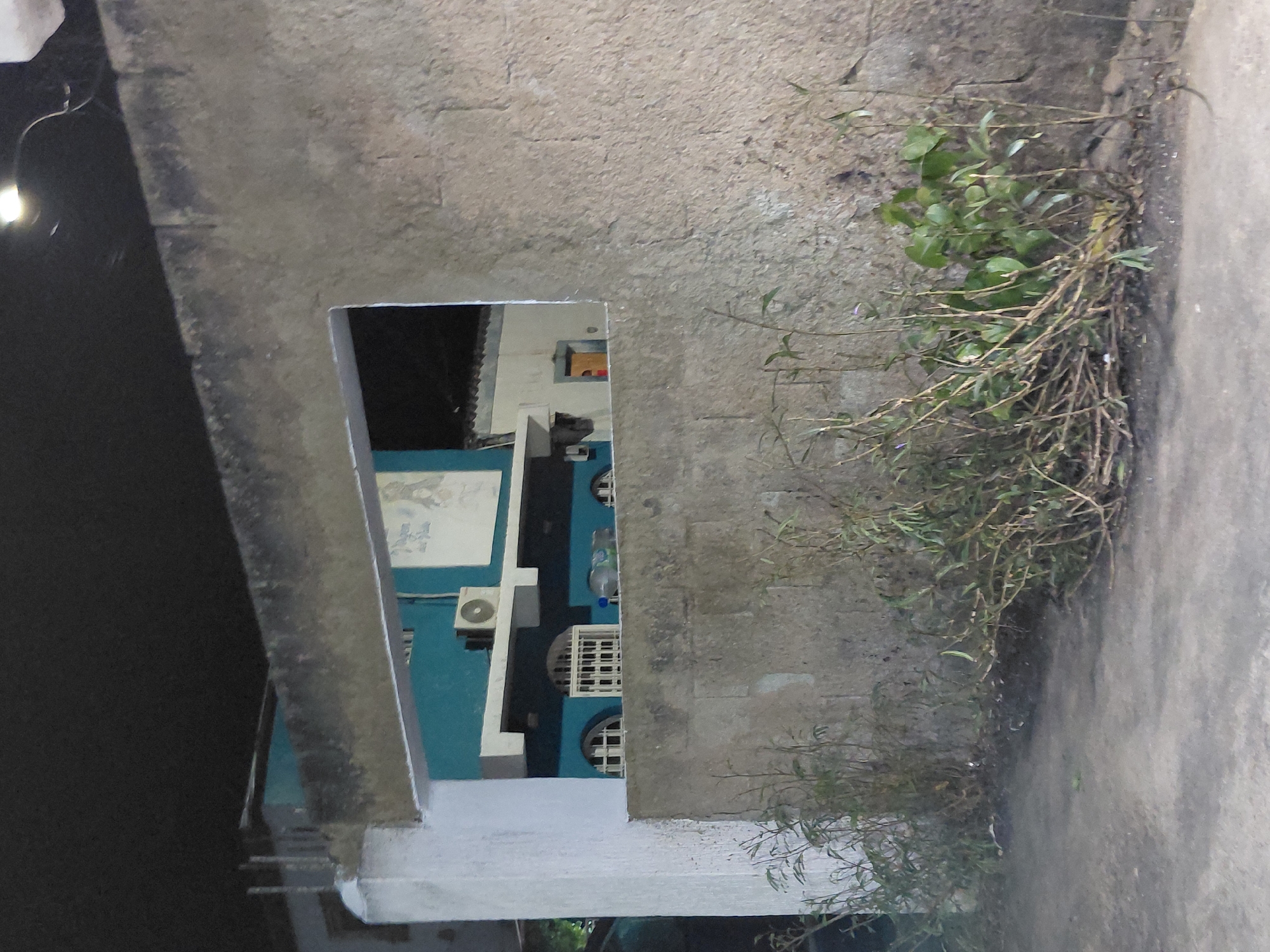 This image shows a view through an opening in a concrete structure, revealing a blue building. The concrete structure is weathered and partially covered with vegetation. The blue building has a white trim and features architectural details such as windows, an air conditioning unit, and possibly a mural or sign. The setting appears to be an outdoor urban environment. The angle suggests it was taken at ground level. It is difficult to confirm the exact location as Santa Ana, Venezuela, without additional information or landmarks.