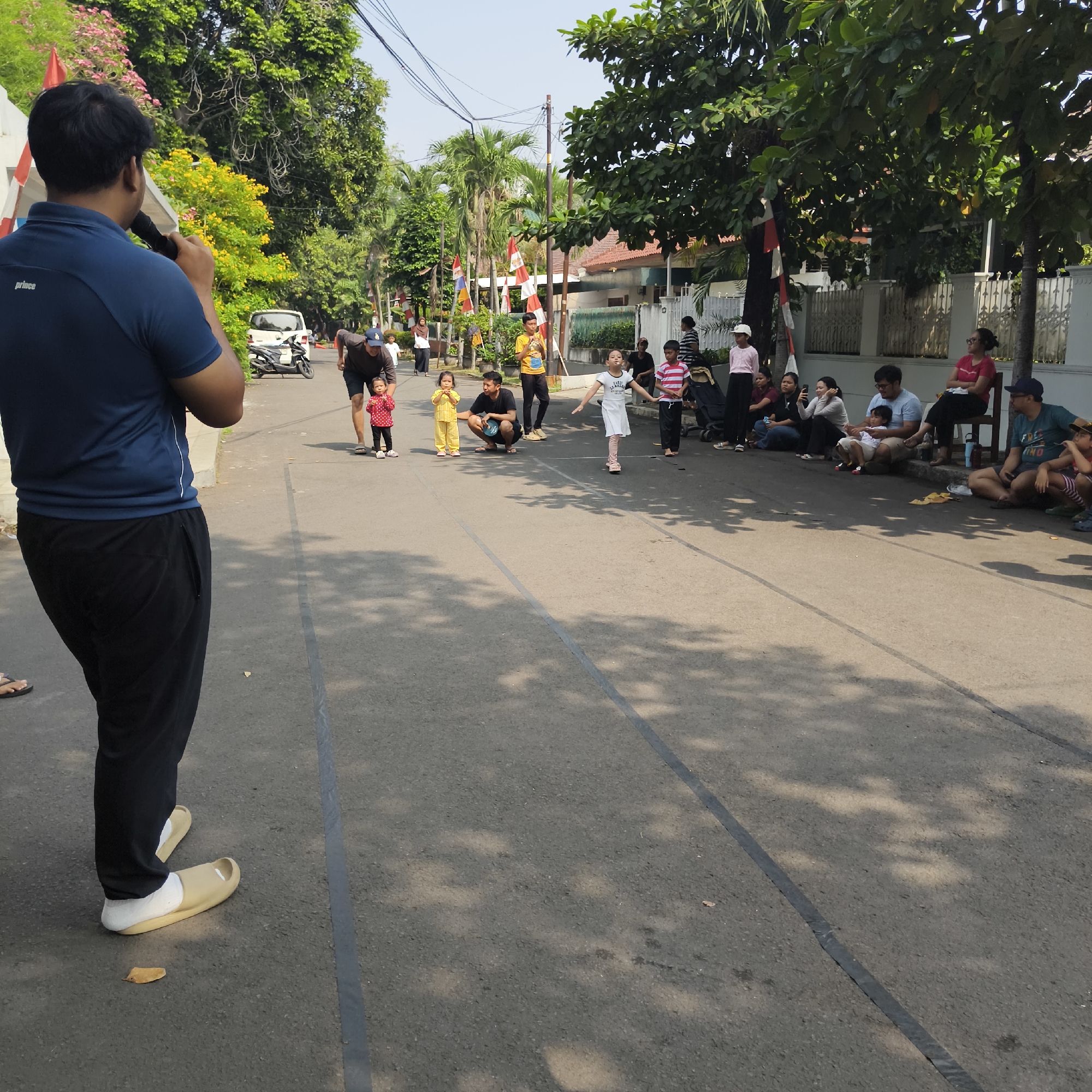 The image depicts an outdoor scene on a street in South Jakarta, Indonesia, where a children's race appears to be taking place. A man in a dark blue polo shirt and black pants, facing away from the camera, holds a microphone, suggesting he is an announcer or facilitator. Several children are participating in or observing the race. One young girl in a white dress is running, with her arms outstretched. Another child in a red and white striped shirt stands nearby, and a younger child in a yellow outfit is also present. Adults are seated along the side of the street, watching the event. A white van and a motorcycle are parked on the side of the road. The street is lined with trees and houses, and Indonesian flags are visible, indicating a festive or celebratory atmosphere, possibly related to a national holiday. The ground is asphalt, with dark lines marking lanes or boundaries for the race. The lighting suggests it is a sunny day.