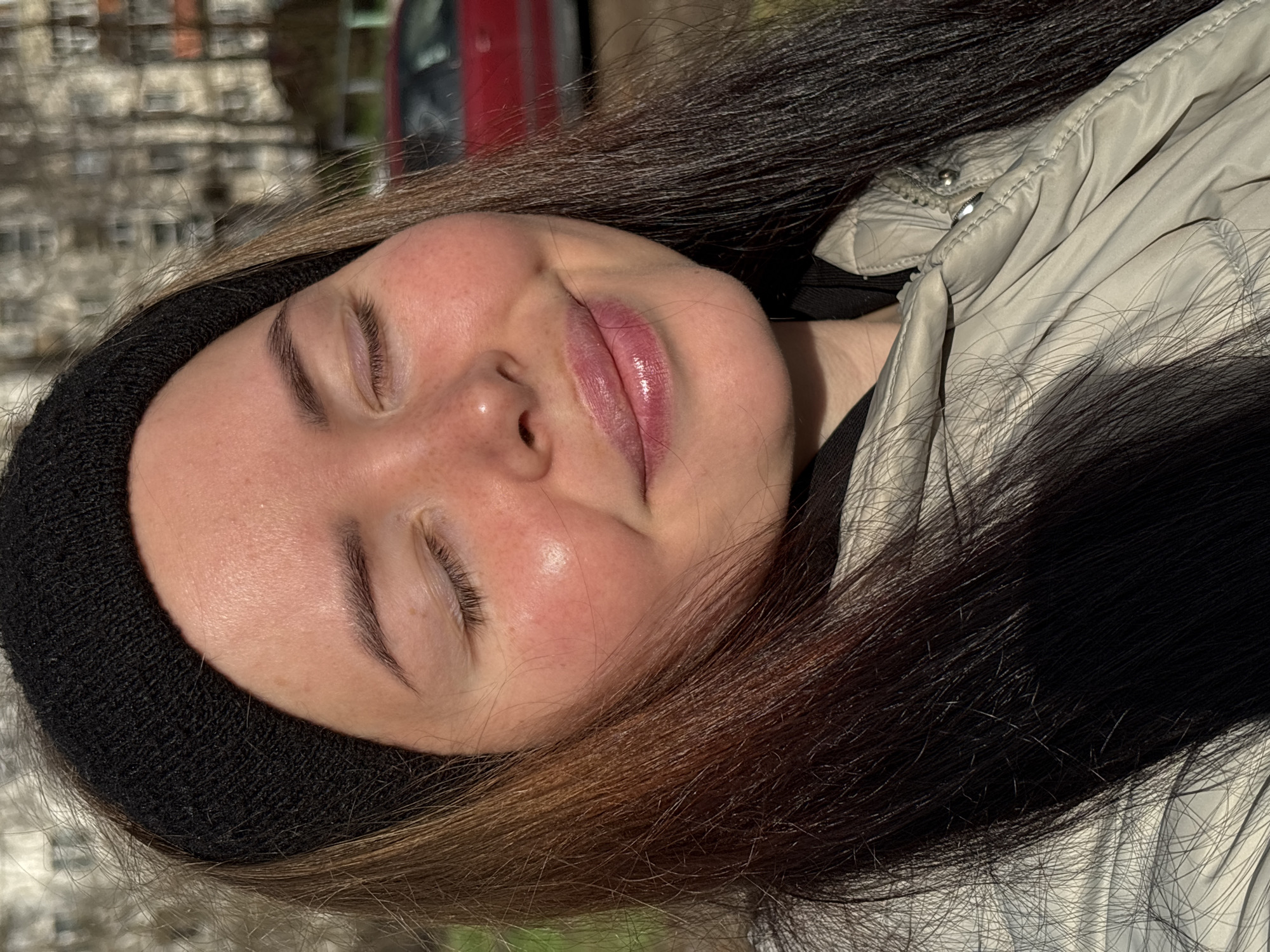 A close-up, rotated portrait captures a young person with long, dark brown hair, featuring lighter brown streaks, flowing around their head and shoulders. The person's eyes are closed, and a gentle smile plays on their lips, which are glossy and pinkish. Their skin shows a natural flush on the cheeks, highlighted by strong sunlight.
They are wearing a black knit beanie that covers their forehead and ears, and a light-colored, possibly cream or beige, puffy jacket. The jacket has visible stitching and buttons, suggesting a casual, warm garment suitable for cooler weather.
The background is softly blurred but reveals elements of an urban environment. A red vehicle is visible on the right side, and behind it, a building with multiple windows can be discerned, indicating a city street or outdoor area. The bright, direct sunlight suggests it is daytime, possibly late morning or afternoon. The overall impression is one of peaceful relaxation, as if the person is enjoying a moment of sunshine. There is no visible text in the image. The location context indicates this scene takes place in Izhevsk, Russia.