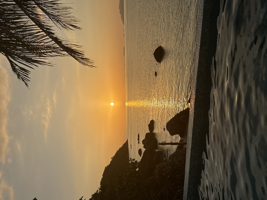 The image captures a sunset over the Andaman Sea in Ban Ko Li Pe, Thailand. The foreground features the dark, rippled surface of a swimming pool, bordered by a tiled edge composed of small, light and dark grey squares. Beyond the pool, the ocean is visible, with a prominent, luminous golden reflection of the sun stretching from the horizon towards the viewer. The sun, a distinct orange-yellow disc, is positioned low over the water in the central-right portion of the frame. To the left, a dark, vegetated landmass, likely an island or hill, slopes into the sea, its base dotted with several dark, irregular rock formations partially submerged in the water. A slender, dark, vertical pole-like object is visible near the shoreline rocks on the left. Faint outlines of distant landmasses are discernible on the right horizon. The sky exhibits a gradient of orange and yellow hues, transitioning to lighter tones with subtle cloud formations near the top. A dark palm frond partially extends into the upper right corner of the frame. No individuals are depicted in the scene.