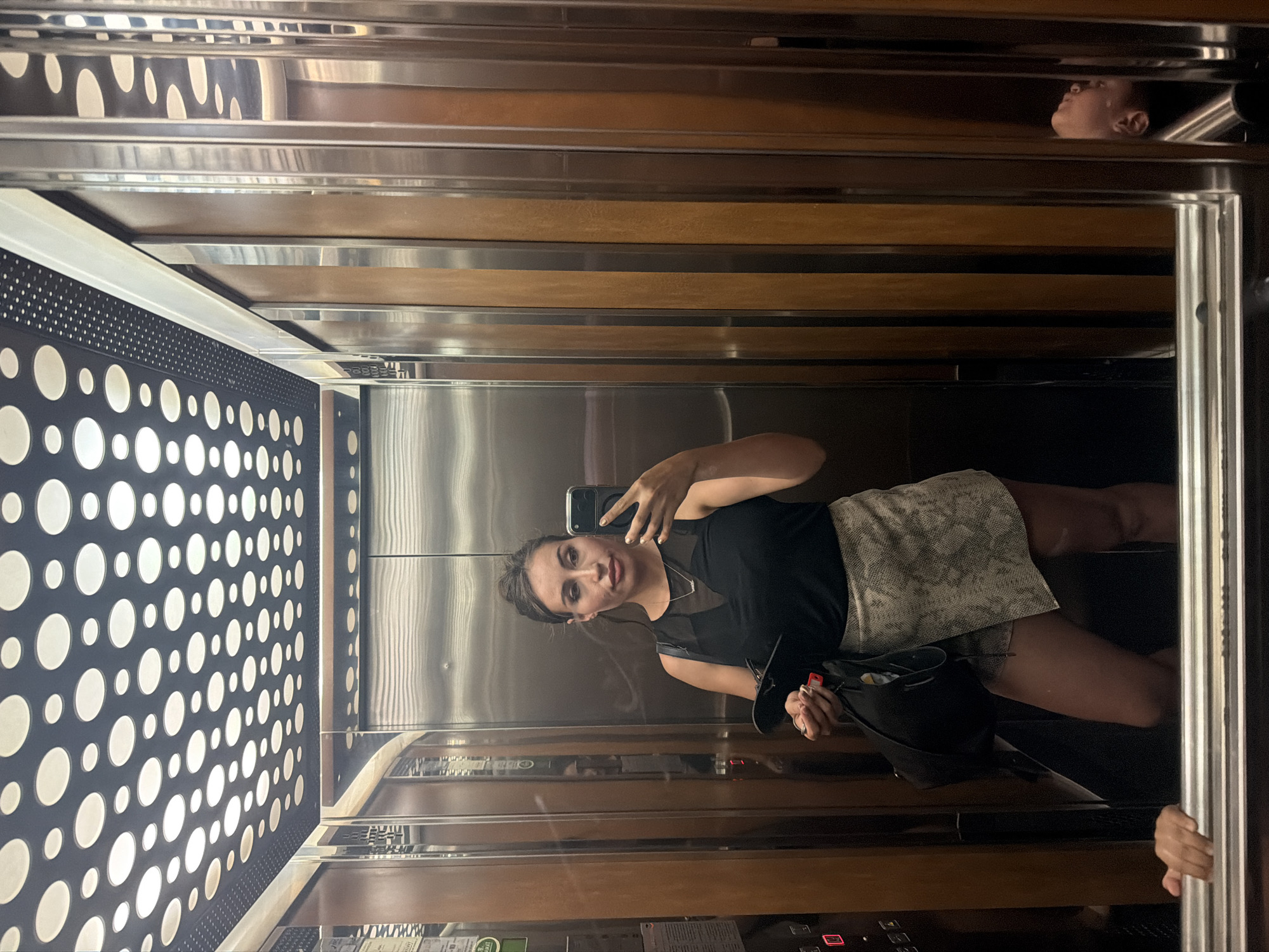 A woman is taking a selfie in an elevator. She is wearing a black top and a patterned skirt. She is holding a bag in her left hand and a phone in her right hand. The elevator has wood-paneled walls and a mirrored ceiling with a circular pattern of lights. There is a reflection of another person in the mirror on the right side of the elevator. The scene appears to be in Tersefanou, Cyprus, though this cannot be definitively confirmed from the image alone.