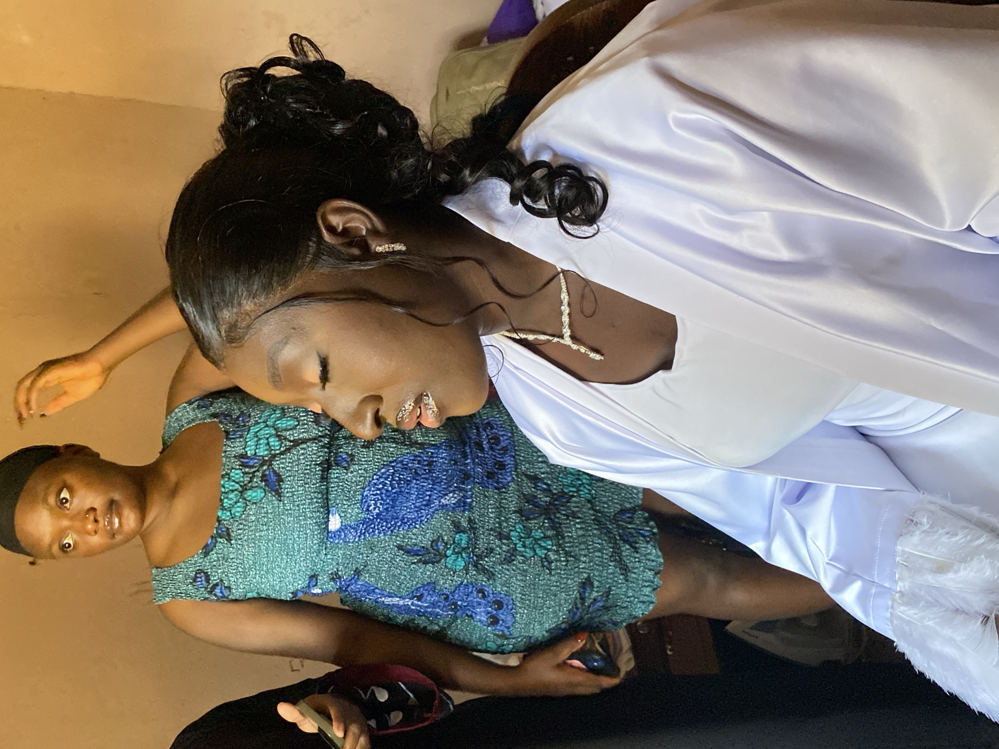 The image shows two women indoors. One woman is wearing a white satin robe with feathered trim and a diamond necklace and earrings. Her hair is styled in an updo with curls. She is leaning on the shoulder of the other woman, who is wearing a green and blue patterned dress and a black head covering. Both women are looking at the camera. The location is Barikin Sabon Gidan Kanat, Nigeria.