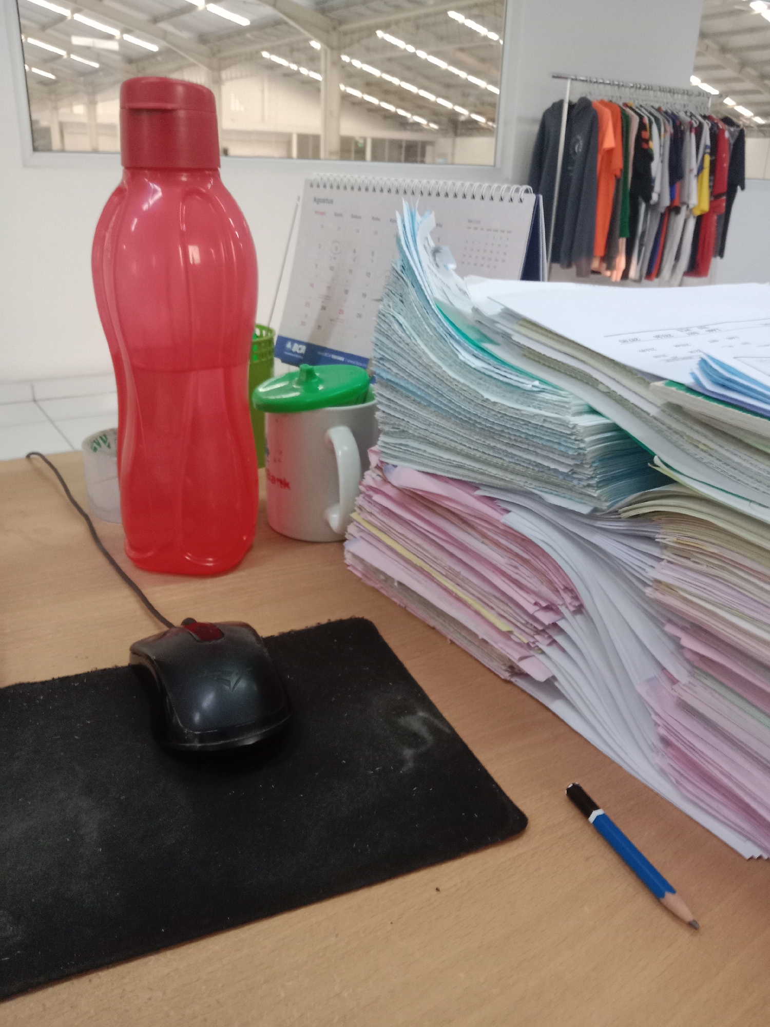 The image depicts a cluttered desk in what appears to be an office or workshop environment. A large, translucent red water bottle stands prominently to the left. To its right, a white mug with a green lid is visible, with a green object, possibly another container or holder, partially obscuring the mug. A stack of papers, fanned out and of varying colors (white, pink, blue, and green), dominates the right side of the frame. A black computer mouse rests on a black mousepad in the foreground. A blue pencil with a black tip lies on the wooden desk surface to the right of the mousepad. In the background, a rack of colorful t-shirts is displayed against a white wall, and a window offers a view of what seems to be a large industrial space with a high ceiling and numerous fluorescent lights. The overall impression is one of an active workspace with an accumulation of daily use items. No specific landmarks or indicators point to the city of Semarang, Indonesia.