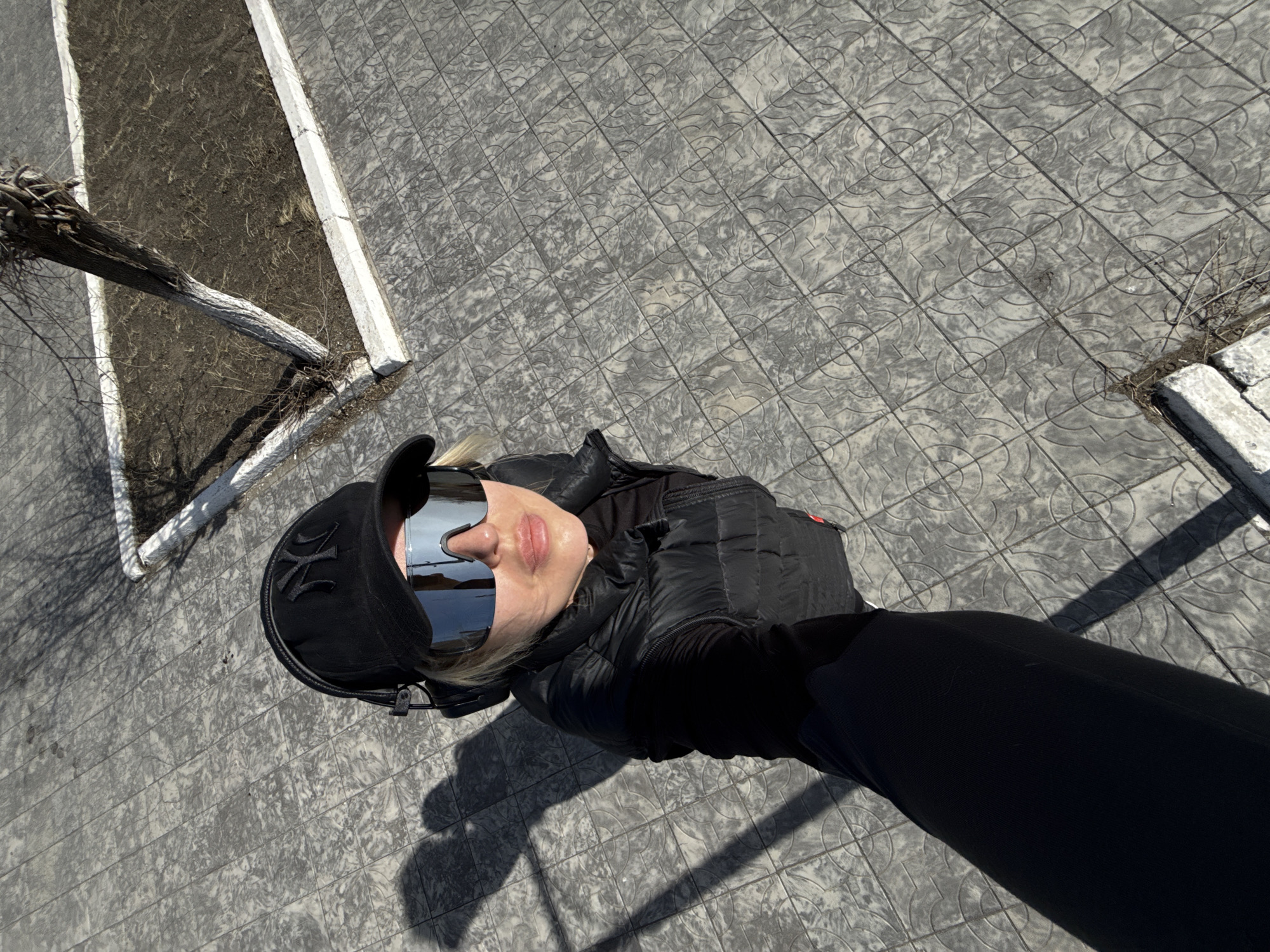 This high-angle shot, taken from above, captures a person looking up at the camera in an outdoor setting in Borzya, Russia. The individual, appearing to be female, is positioned centrally in the frame, surrounded by patterned paving stones.
The person is dressed in dark attire, suggesting cool or cold weather. They wear a black cap with a prominent, embroidered logo resembling "NY" (New York Yankees) on the front. Large, highly reflective, futuristic-style sunglasses cover most of their face, mirroring the sky and surroundings. Their lips are visible, appearing full and a light pink shade. The person is also wearing a black puffer jacket or vest over a long-sleeved black garment and black gloves. Blonde hair peeks out from under the cap.
The ground beneath the person is covered with grey paving stones, arranged in a grid pattern, each tile featuring a debossed circular or geometric design. To the upper left and right of the frame, there's a narrow strip of barren, dry soil or ground, bordered by white concrete curbs. A portion of a gnarled, possibly cut or severely pruned, tree trunk with some bare branches extends from this strip of ground.
The strong, distinct shadows cast by the person and the tree indicate bright sunshine and a clear sky, suggesting it is daytime, possibly late morning or early afternoon. The overall mood is neutral, with the person's expression largely obscured by the sunglasses.