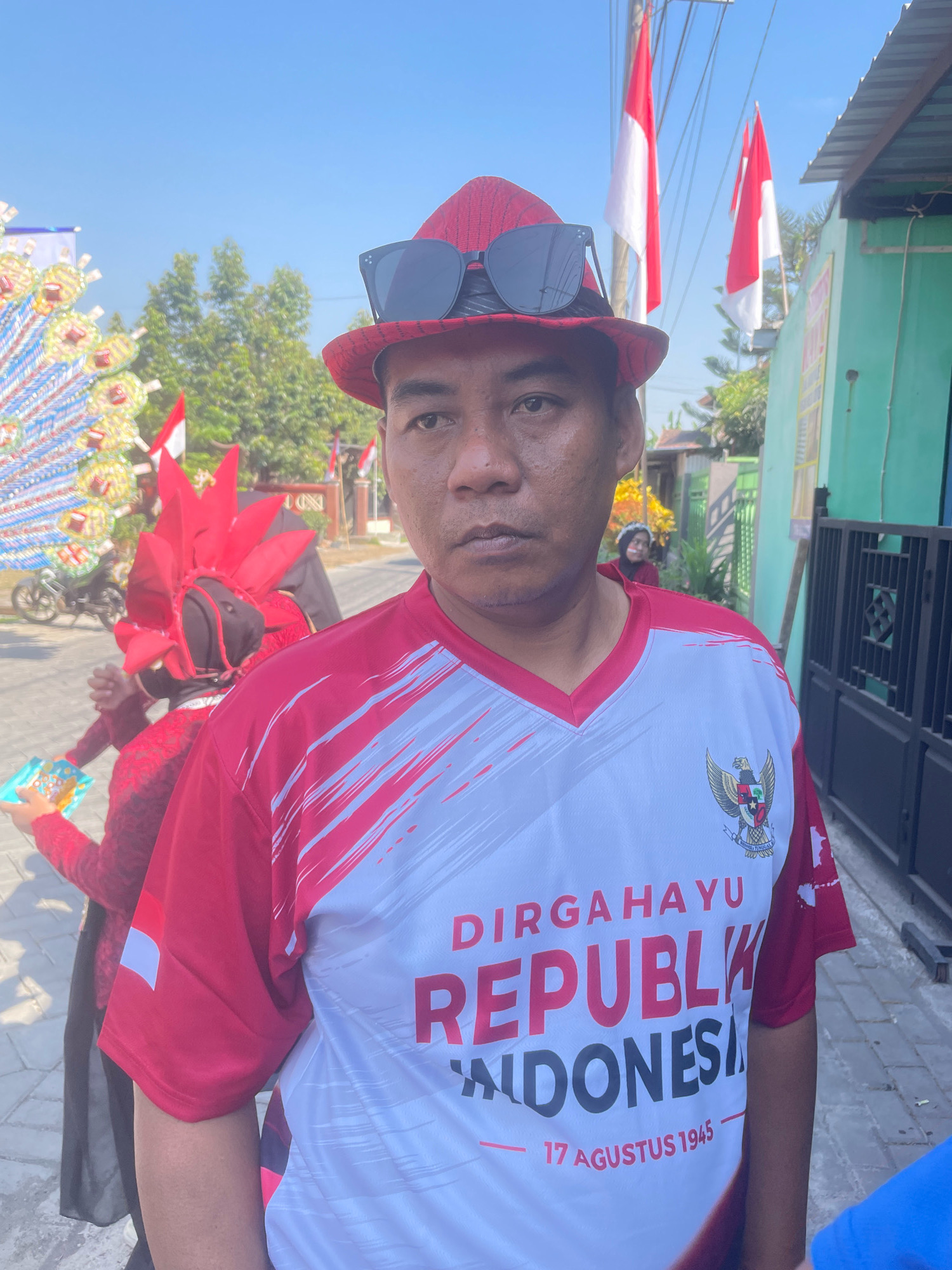 In Kebon Waris, Indonesia, a man wearing a red and white patterned shirt with "DIRGAHAYU REPUBLIK INDONESIA 17 AGUSTUS 1945" printed on it stands facing the camera. He wears a red fedora with sunglasses perched on top. In the background, two Indonesian flags fly from poles. To the left, a person in a red costume with a spiky headpiece and black veil is visible, holding a colorful object. Another person wearing a red and white headscarf is further in the background. The environment appears to be outdoors on a sunny day, with trees and buildings visible.