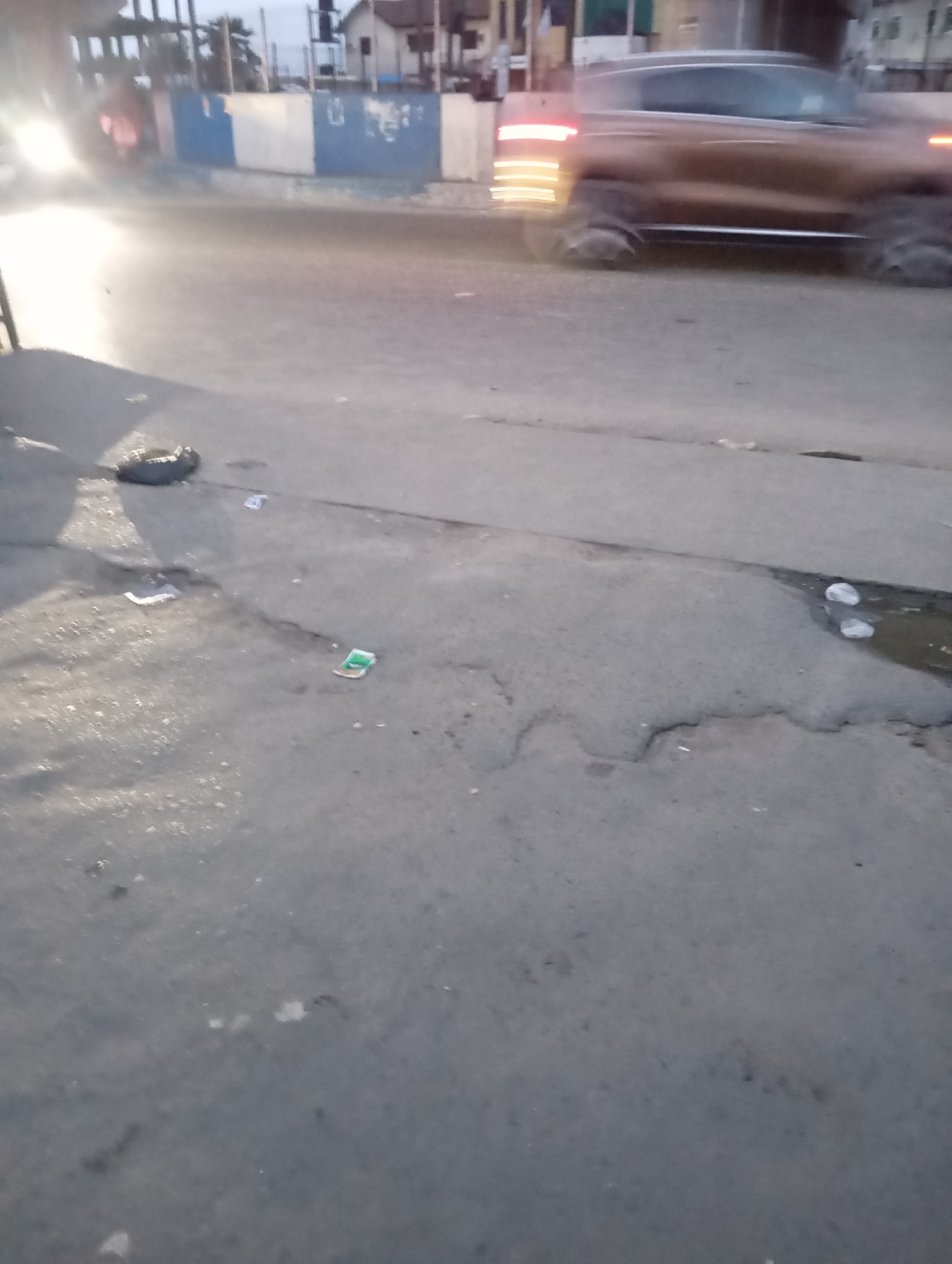 A street scene in Port Harcourt, Nigeria, features a blurred image of a brown SUV driving on a paved road. The vehicle's taillights are illuminated and appear streaked due to motion blur. The road surface is asphalt, with visible cracks and patches. Scattered debris, including what appears to be a plastic bag, a crumpled piece of paper, and some small white objects, lies on the roadside. A small puddle with two white circular objects in it is visible on the right side of the frame. In the background, a low building with a blue and white patterned wall can be seen, along with structures that suggest urban infrastructure. The lighting indicates it is likely dusk or dawn, with artificial lights from the vehicle and possibly streetlights contributing to the scene's illumination.