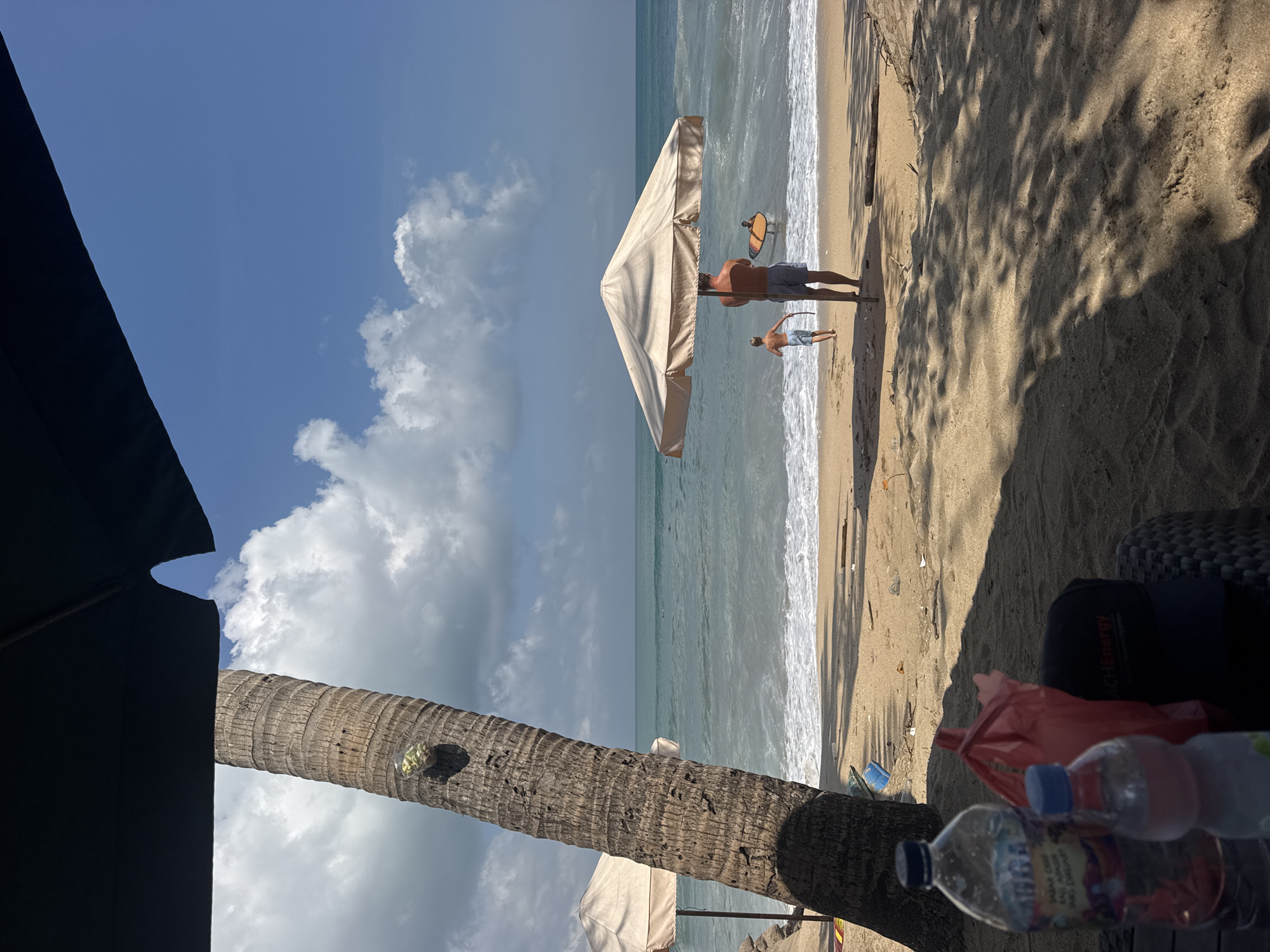The image captures a sunny day at a beach in Kuta, Indonesia. In the foreground, partially obscured by a large black umbrella and the trunk of a coconut palm tree, are two plastic water bottles and a red plastic bag on the sand. The sand is tan and textured, with dappled sunlight and shadows cast by foliage.
The middle ground features the shoreline with gentle waves breaking on the sand. Two individuals are visible near the water. A man, wearing dark shorts, stands facing away from the camera, holding a large beach umbrella. Beside him, a young boy, wearing light-colored shorts, runs towards the ocean. Further out in the water, another person is visible with what appears to be a surfboard. Two beige beach umbrellas stand further down the beach.
The background consists of the turquoise ocean stretching to the horizon, meeting a clear blue sky dotted with white, fluffy clouds. The overall atmosphere is one of a relaxed beach day.