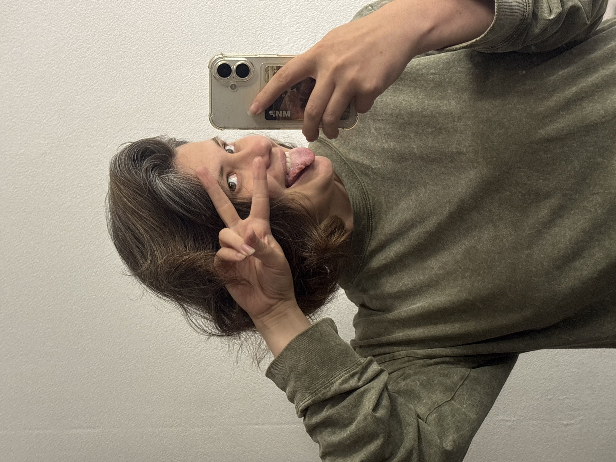 A person is taking a mirror selfie. They are wearing a green sweatshirt and have brown hair with some gray streaks. The person is sticking out their tongue and making a peace sign with their fingers. They are holding a smartphone in their hand to take the picture. The background appears to be a wall. The lighting suggests it could be daytime indoors.