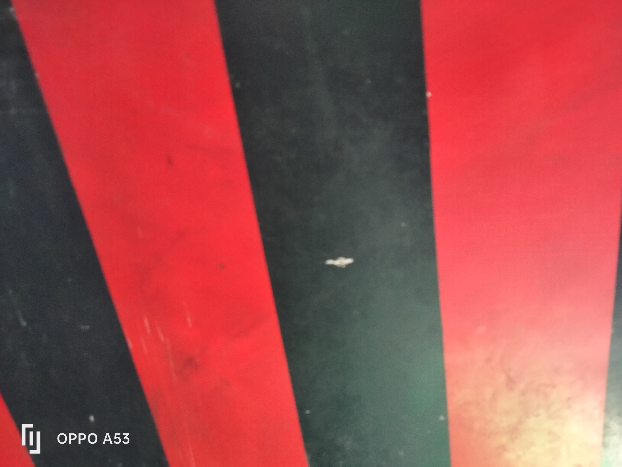 The image displays a close-up view of a surface with alternating red and black stripes. The stripes appear to be painted or printed onto a flat material. There is a small, light-colored mark in the center of the frame, possibly a speck of dust or a scratch. In the bottom left corner, a white logo and the text "OPPO A53" are visible, indicating the device used to capture the image. The image does not provide any information about the city of Denpasar, Indonesia, or any contextual details of a crime scene.
