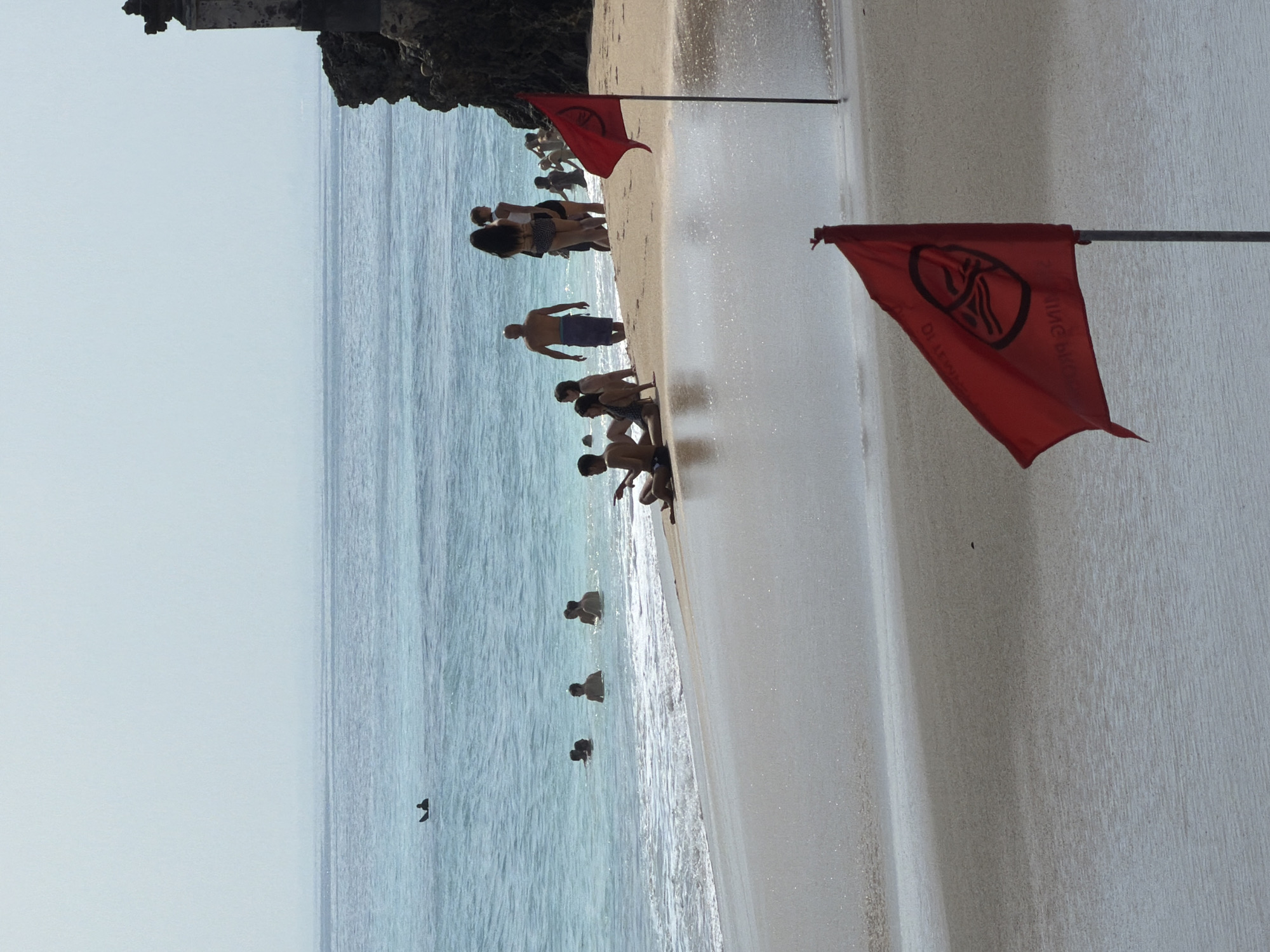 The image depicts a beach scene in Kuta Selatan, Indonesia, characterized by a shoreline with gentle waves and a hazy sky. In the foreground, two red flags are prominently displayed, one in sharp focus and the other slightly out of focus. These flags bear a symbol which is commonly associated with warnings or restrictions. Several individuals are visible in the water, either swimming or wading. Further down the beach, a group of people are gathered, some sitting and others standing. A rocky outcrop with a man-made structure on top is visible on the right side of the frame, casting a shadow on the beach. The overall atmosphere suggests a tropical setting with moderate human activity.